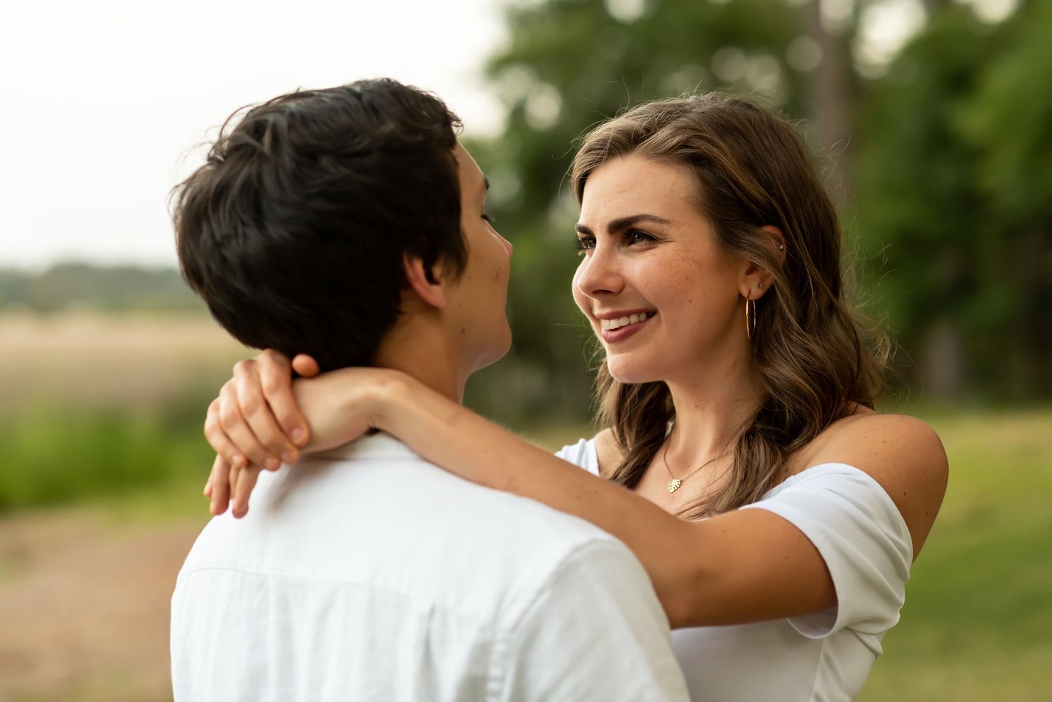A romantic couple embraces outdoors while smiling at each other on a sunny day with a natural landscape in the background.
