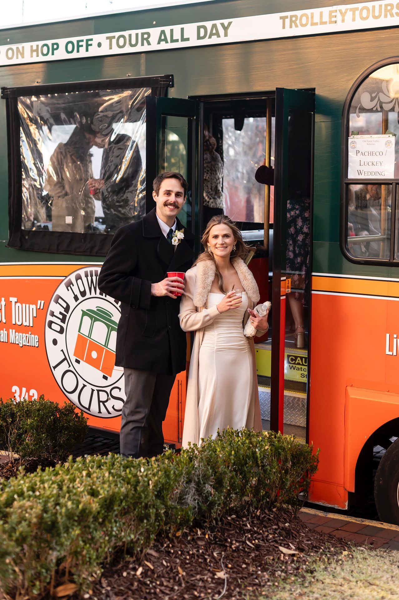 A bride and groom pose together next to an orange trolley tour bus on their wedding day.