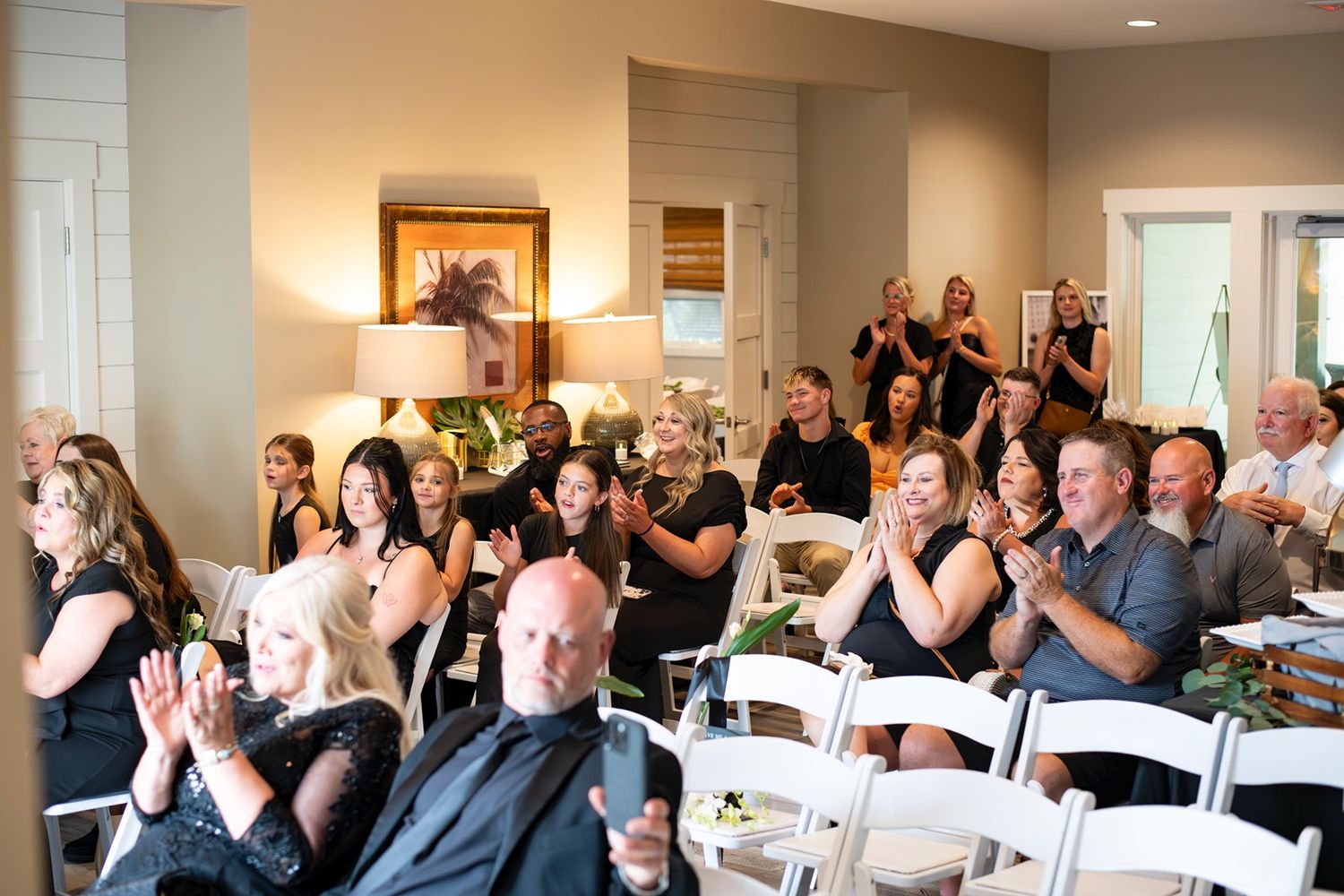 Wedding guests seated in rows of white chairs applaud during an indoor reception celebration.