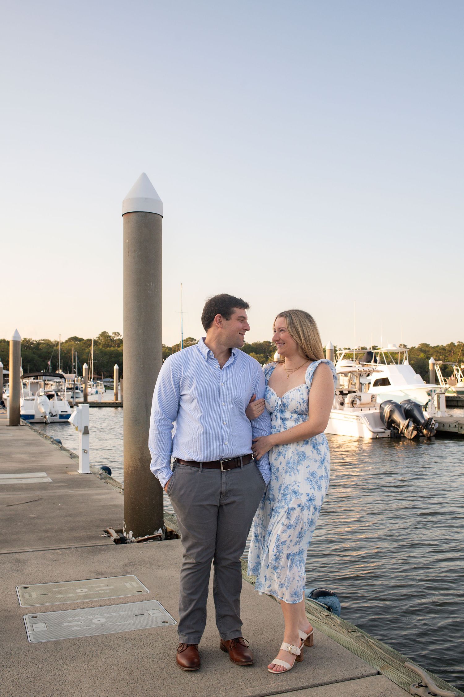A couple shares romantic moments along a marina dock at sunset with boats in the background.