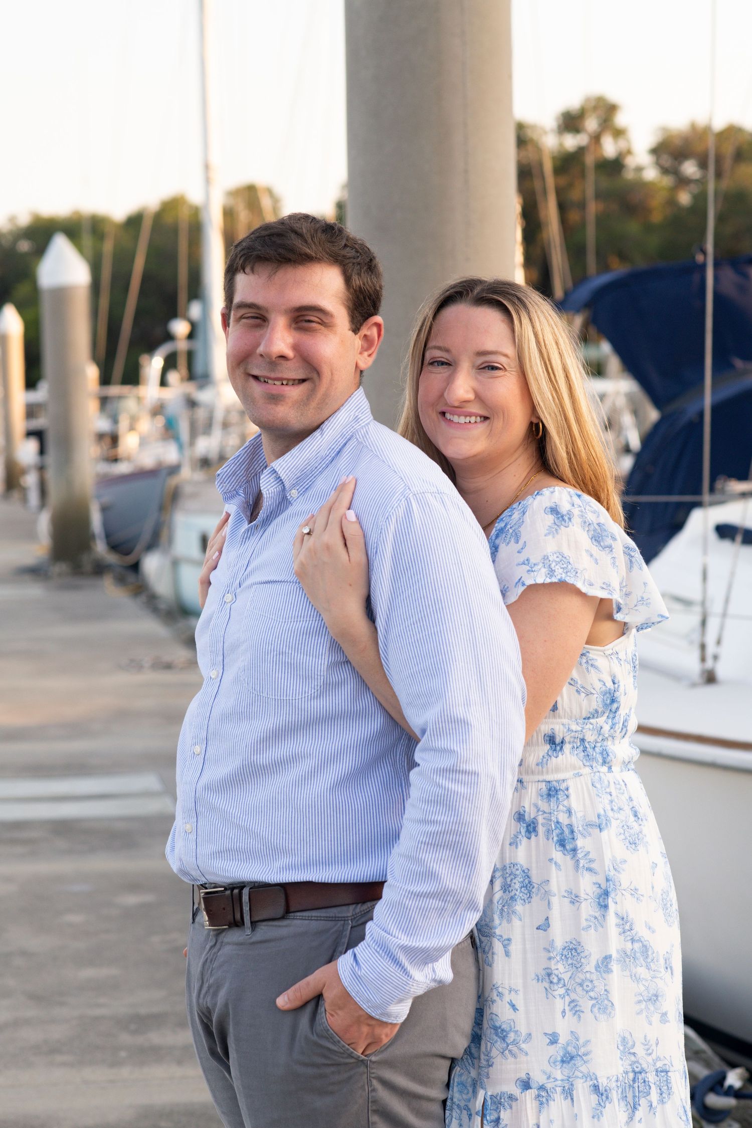 A couple in coordinating blue and white outfits smiles together at a marina with boats and pillars in the background.