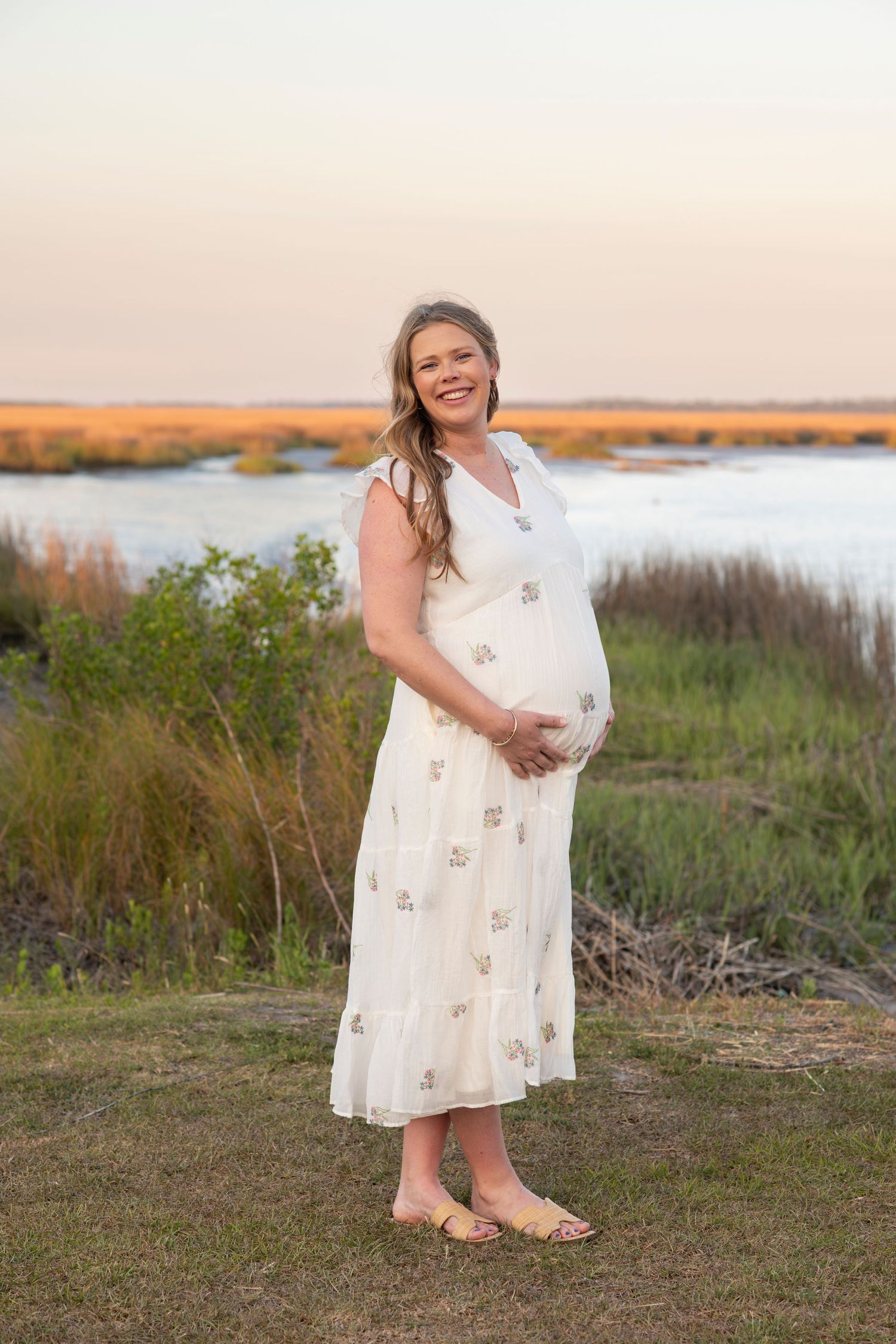 Happy pregnant woman in white dress cradling bump at golden hour by marsh waterway.