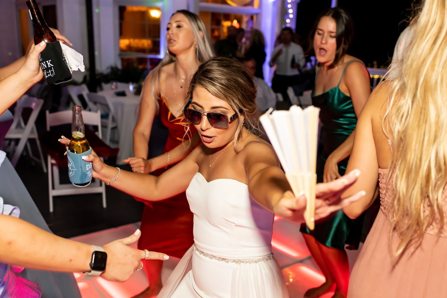 Bride in white dress dancing with bridesmaids in colorful gowns at a lively wedding reception party.