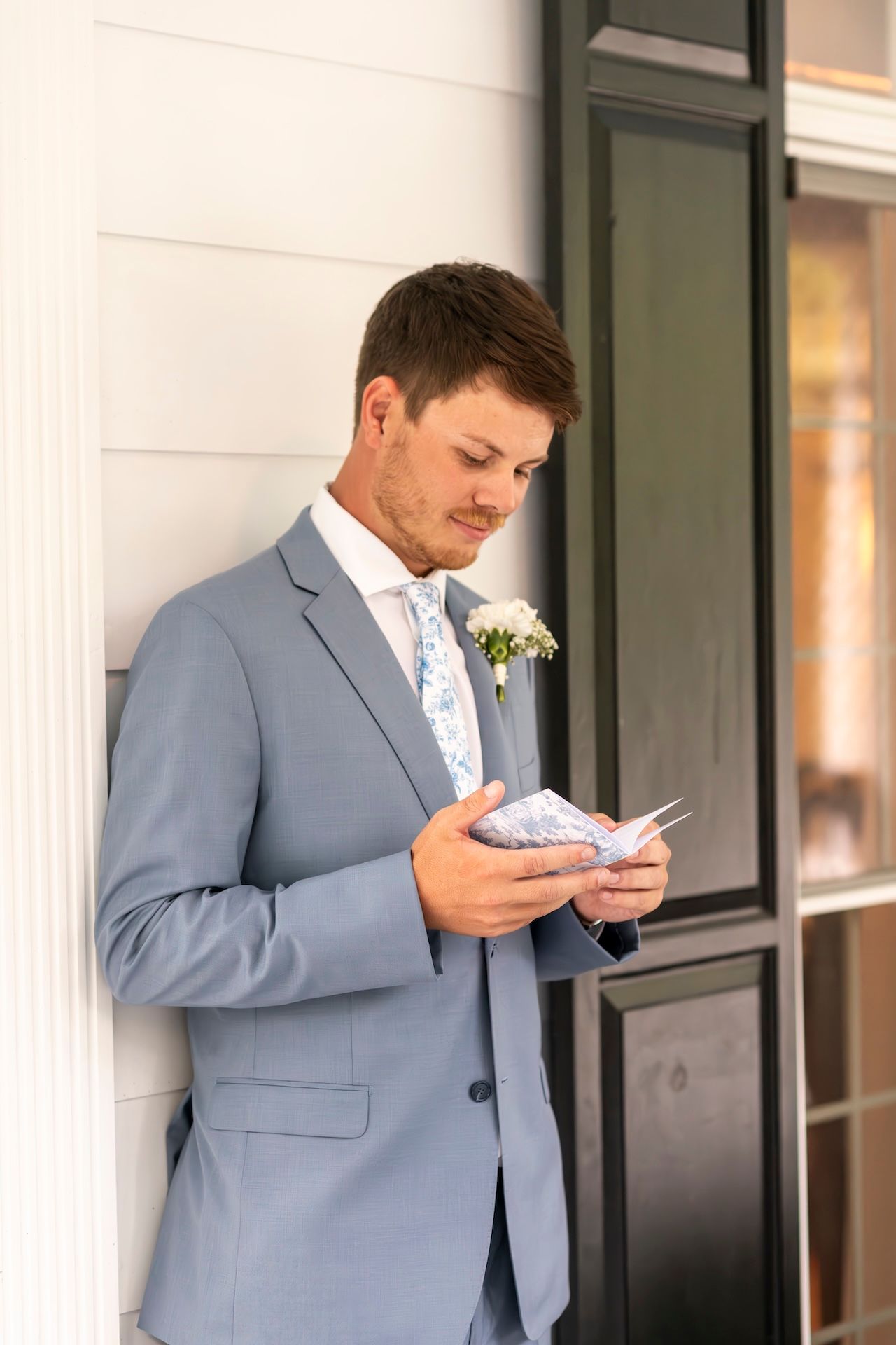 Groom in light blue suit with floral boutonniere reads a note before wedding ceremony outdoors.