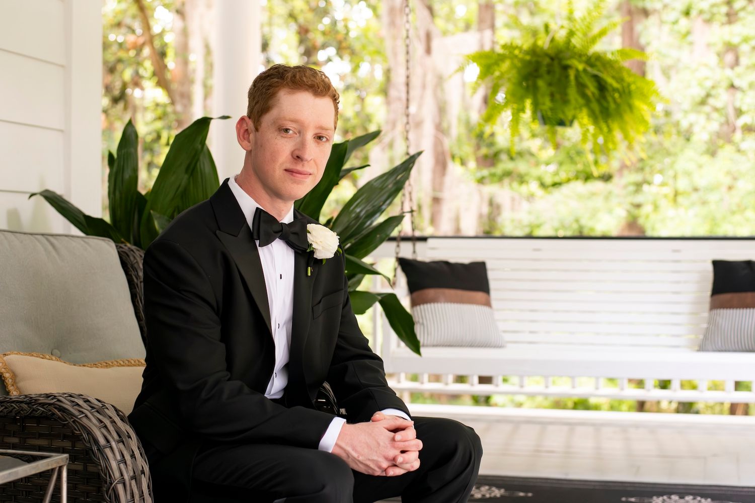 Young man in black tuxedo with white boutonniere sits on porch, posing for formal portrait.