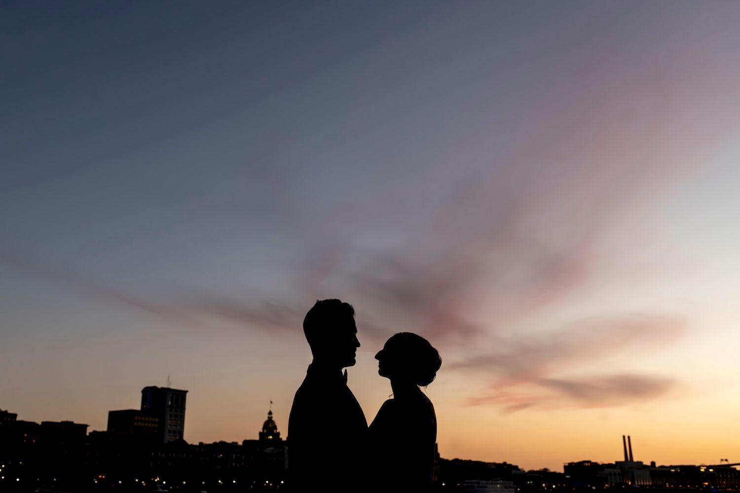 Silhouette of a couple facing each other against a vibrant sunset sky with a city skyline in the background.