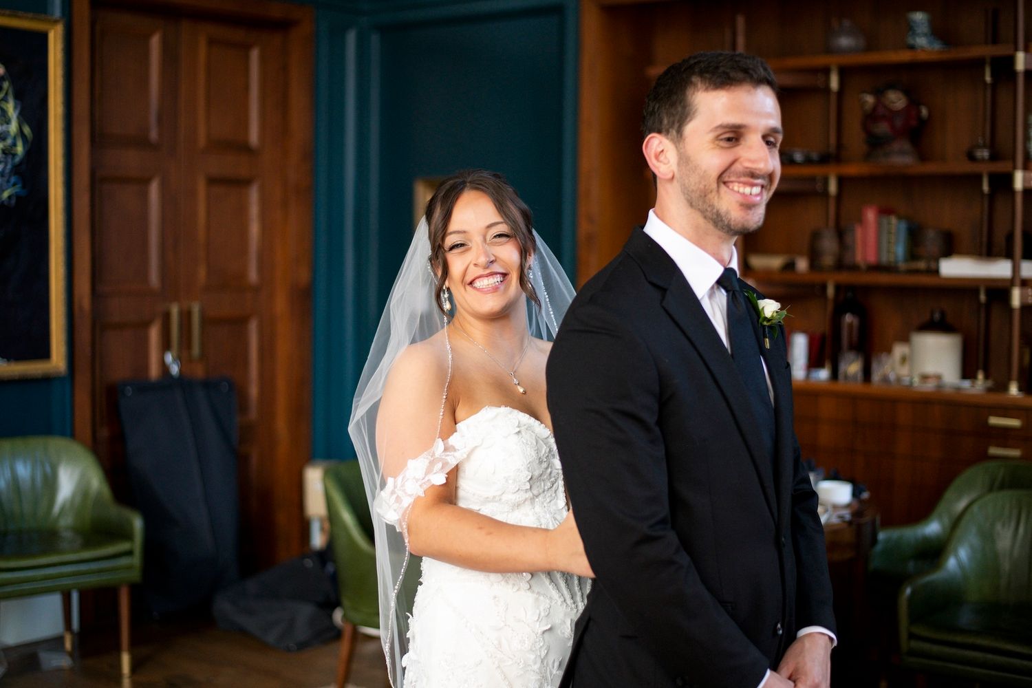 A couple in wedding attire walks together and shares a laugh in an elegant wood-paneled room.