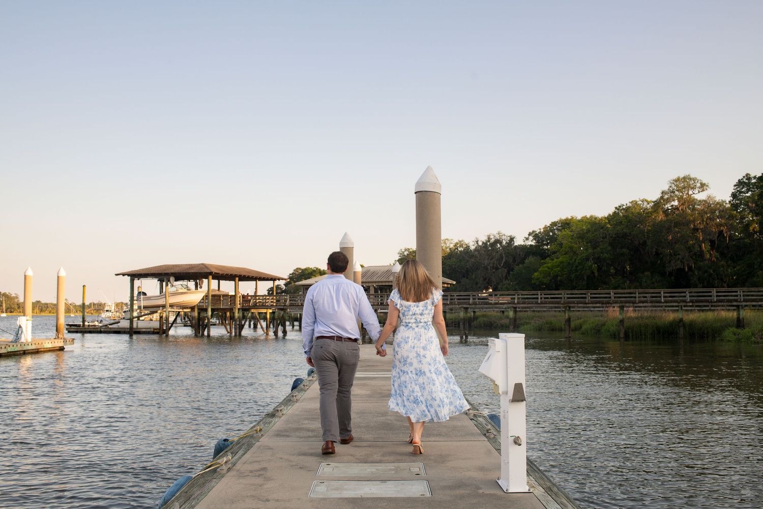 Newlyweds walk along a wooden dock at sunset with boats and water views in the background.