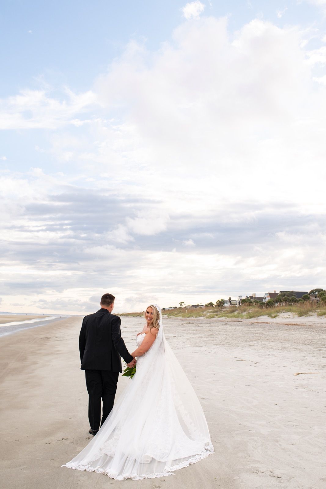 A couple in wedding attire walks along a serene beach shoreline with cloudy skies at sunset.
