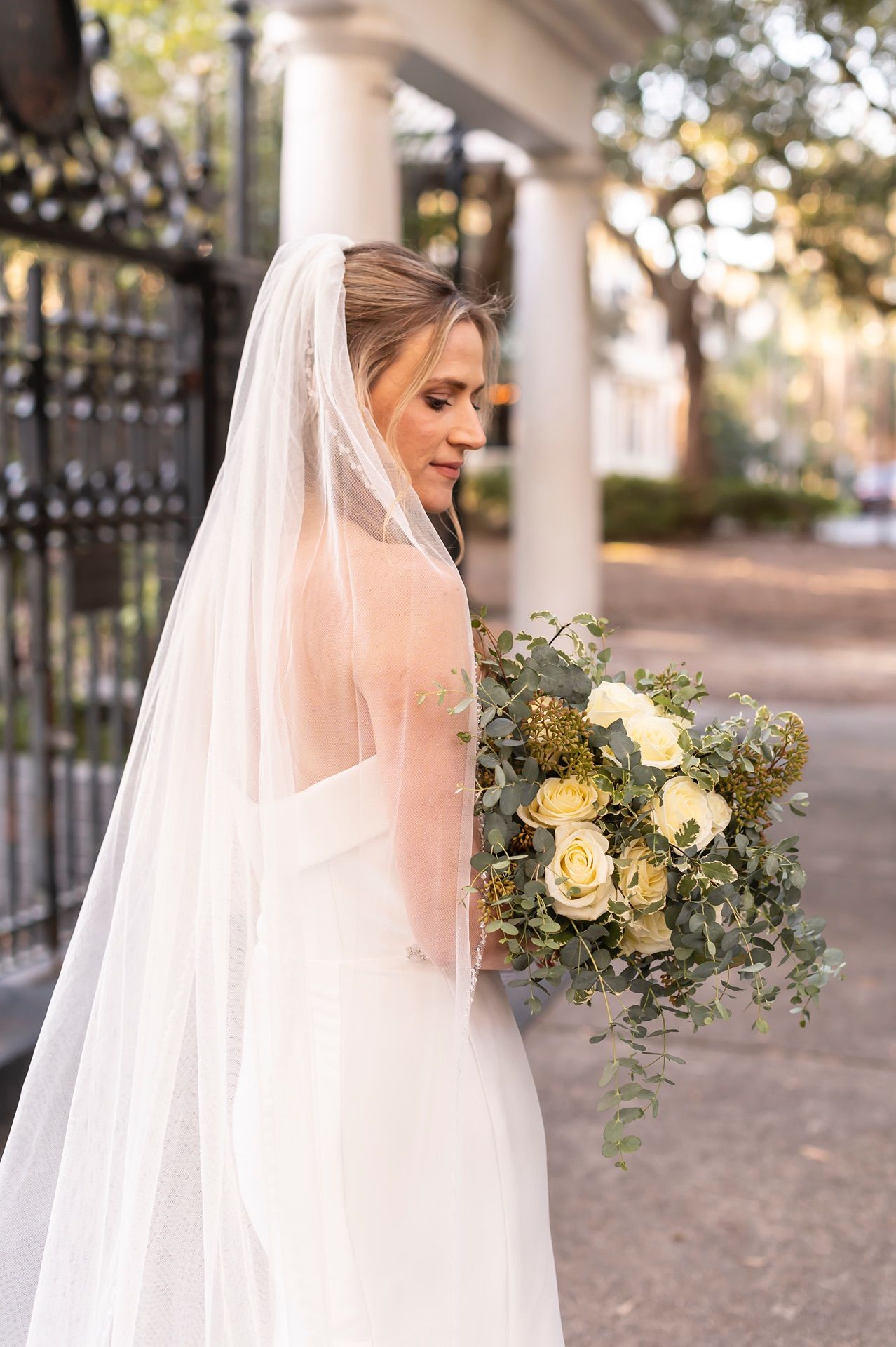 Bride in traditional white dress posing with an elegant bouquet of cream roses and greenery against white columns.