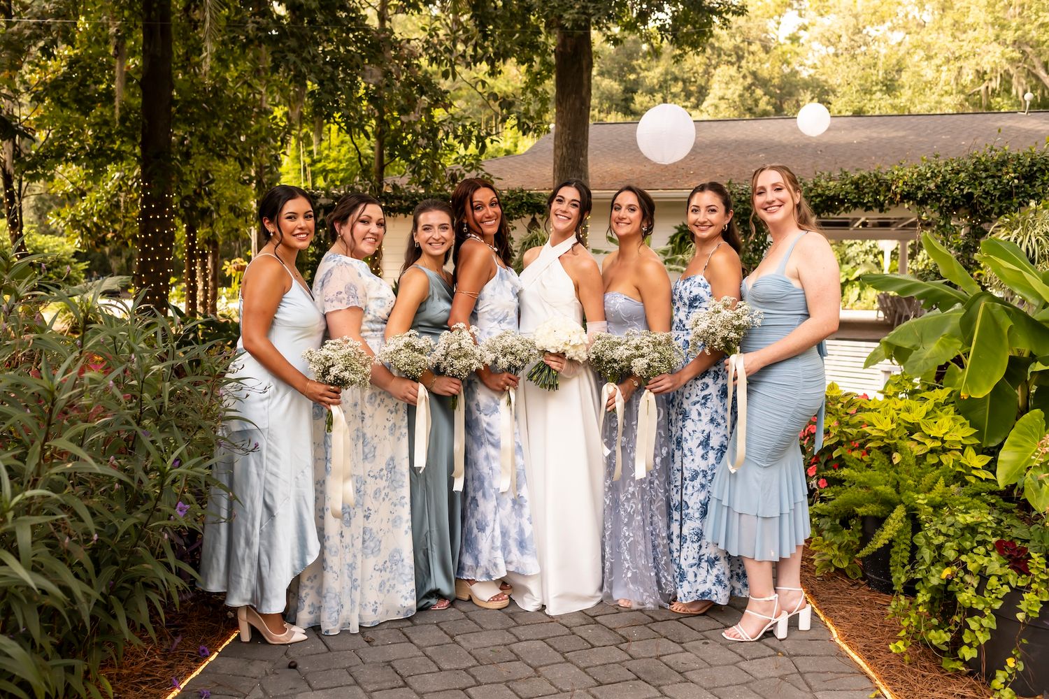 A group in blue and white dresses poses together holding floral bouquets in a lush garden setting.