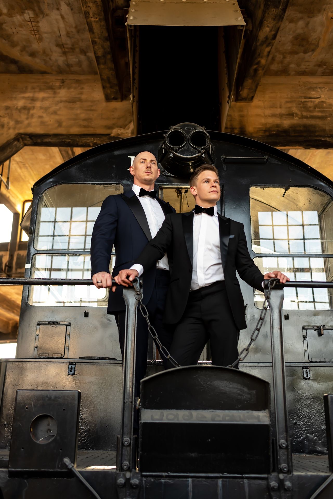 Two men in tuxedos sit atop vintage railroad equipment in an industrial setting with dramatic lighting.