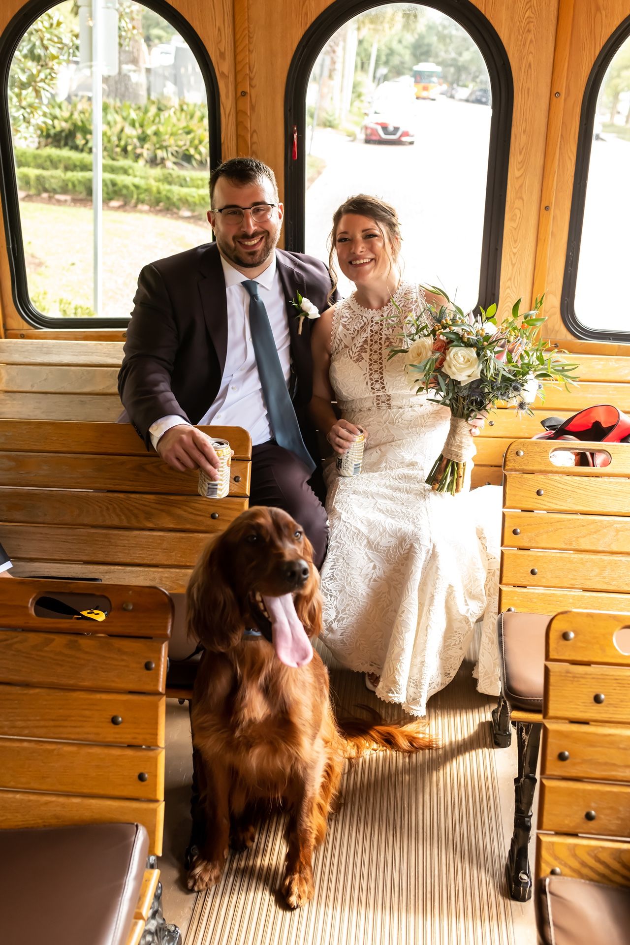 A couple sits happily in a vintage wooden trolley car with their brown dog on their celebratory wedding day.