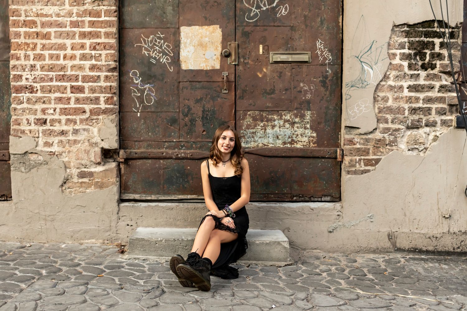 Someone in black attire sits on cobblestone ground against rustic brick wall with weathered metal door.