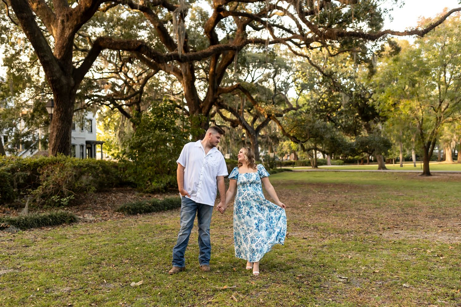 A couple strolls through a peaceful park with Spanish moss-draped oak trees in the background.