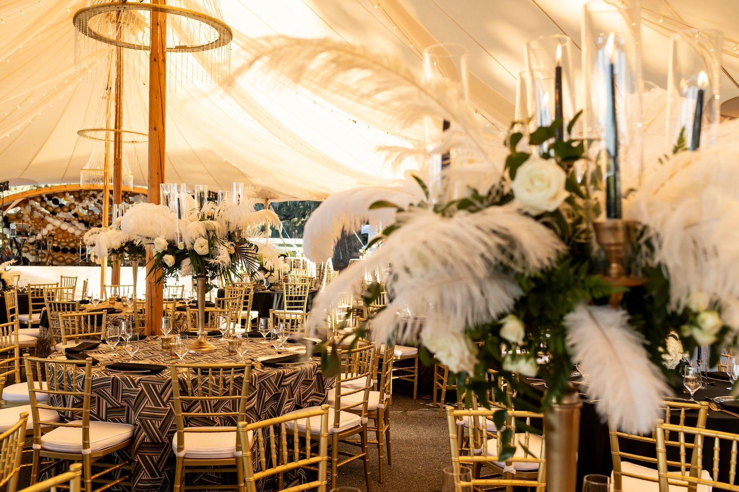 Elegant wedding reception decor featuring tall gold candelabras and white feather centerpieces under a draped tent ceiling.
