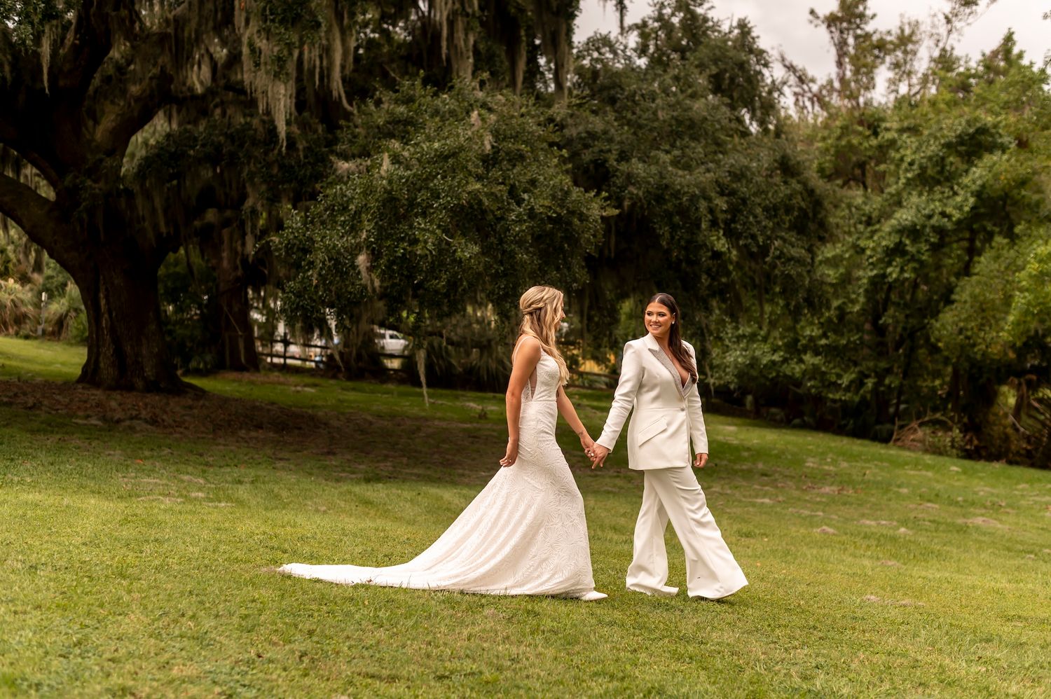 Two people in white attire walk hand in hand across a lush green lawn with Spanish moss draped trees in the background.