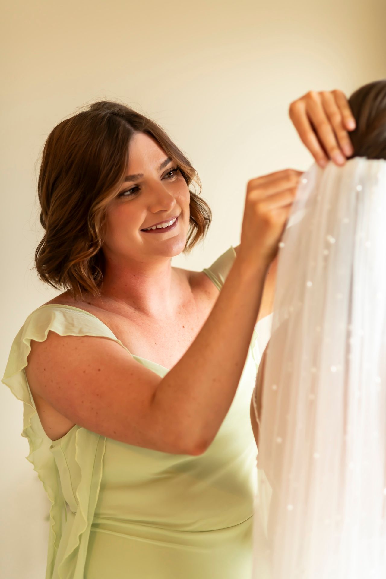 Woman in light green dress adjusts sheer white curtains in a warm-lit room.