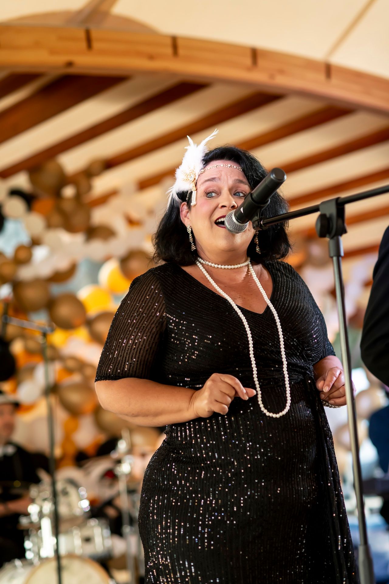 A performer in a sparkling black dress and pearl necklace sings passionately into a microphone on stage with wooden beams above.