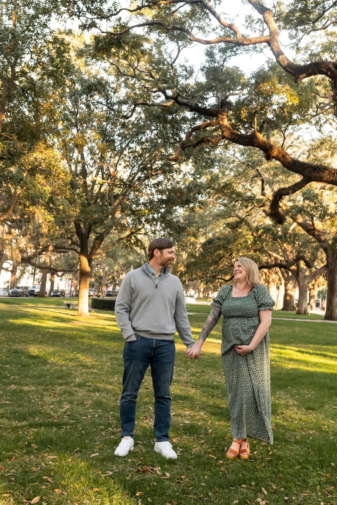 A couple holding hands while standing together on a grassy park area with oak trees during golden hour.