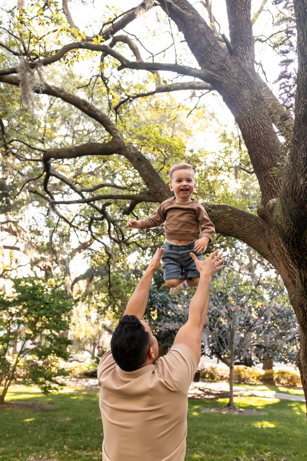 A parent in a beige shirt lifts up a small child wearing a brown shirt next to a large tree in a sunny park.