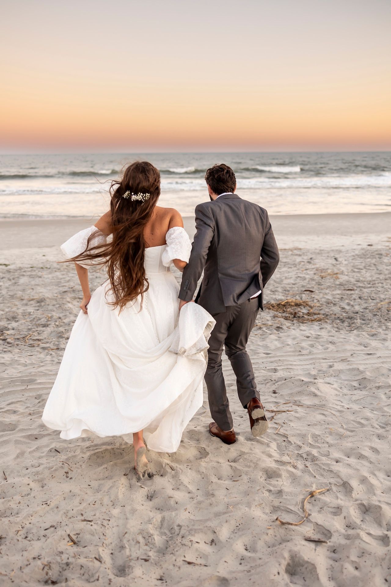 A couple on a beach at sunset with long white wedding dress flowing in the breeze as they walk on the sand.
