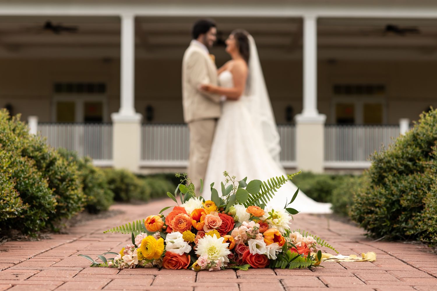 A wedding bouquet of orange and white flowers lies on brick pathway with couple standing in soft focus behind pillared porch.