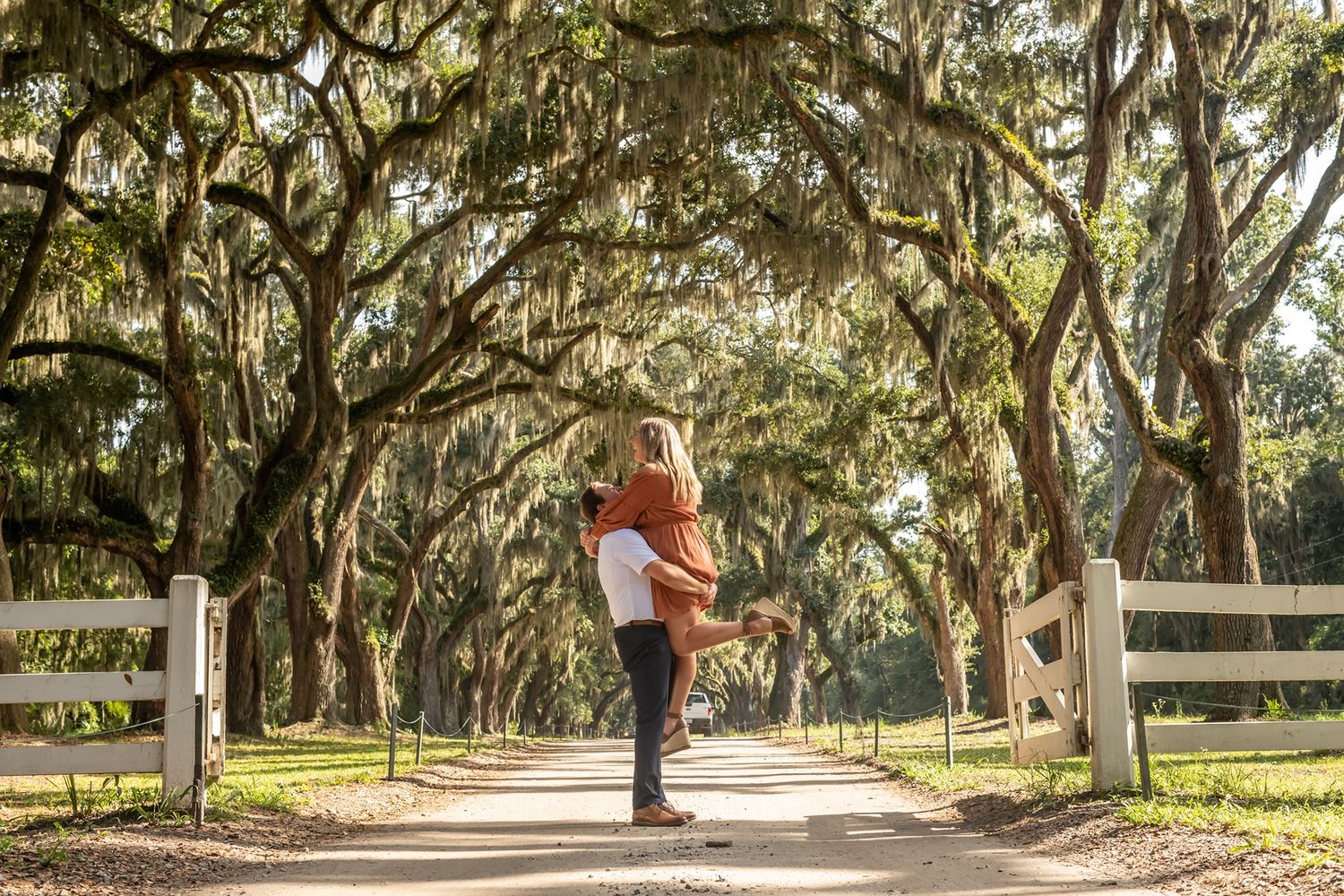 A couple embraces under a canopy of Spanish moss draped oak trees on a fence-lined path in the American South.