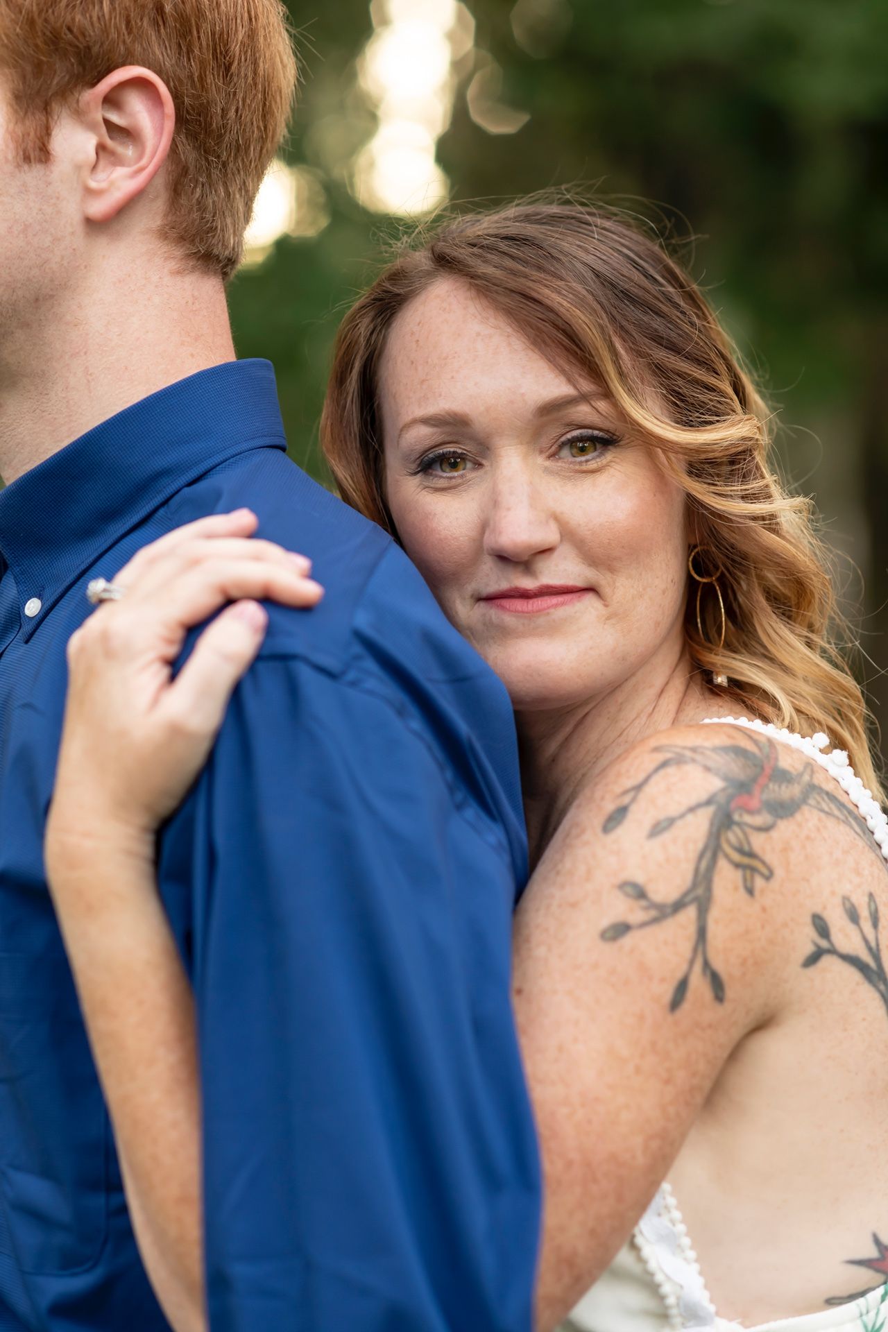 Romantic wedding couple embraces as bride with shoulder tattoo and white dress rests against groom in navy suit.