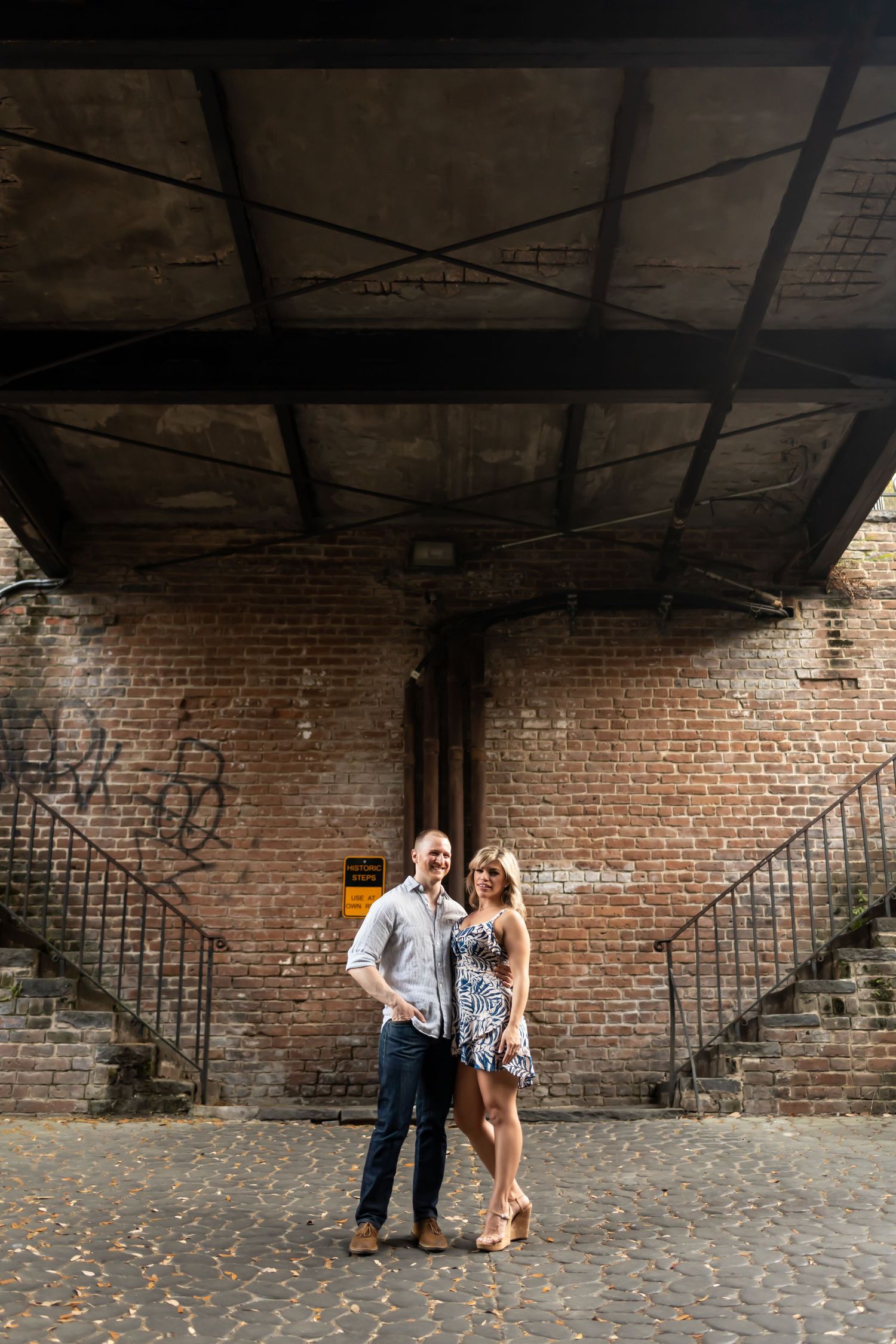 A couple shares a romantic moment against an industrial brick wall backdrop in an urban setting.