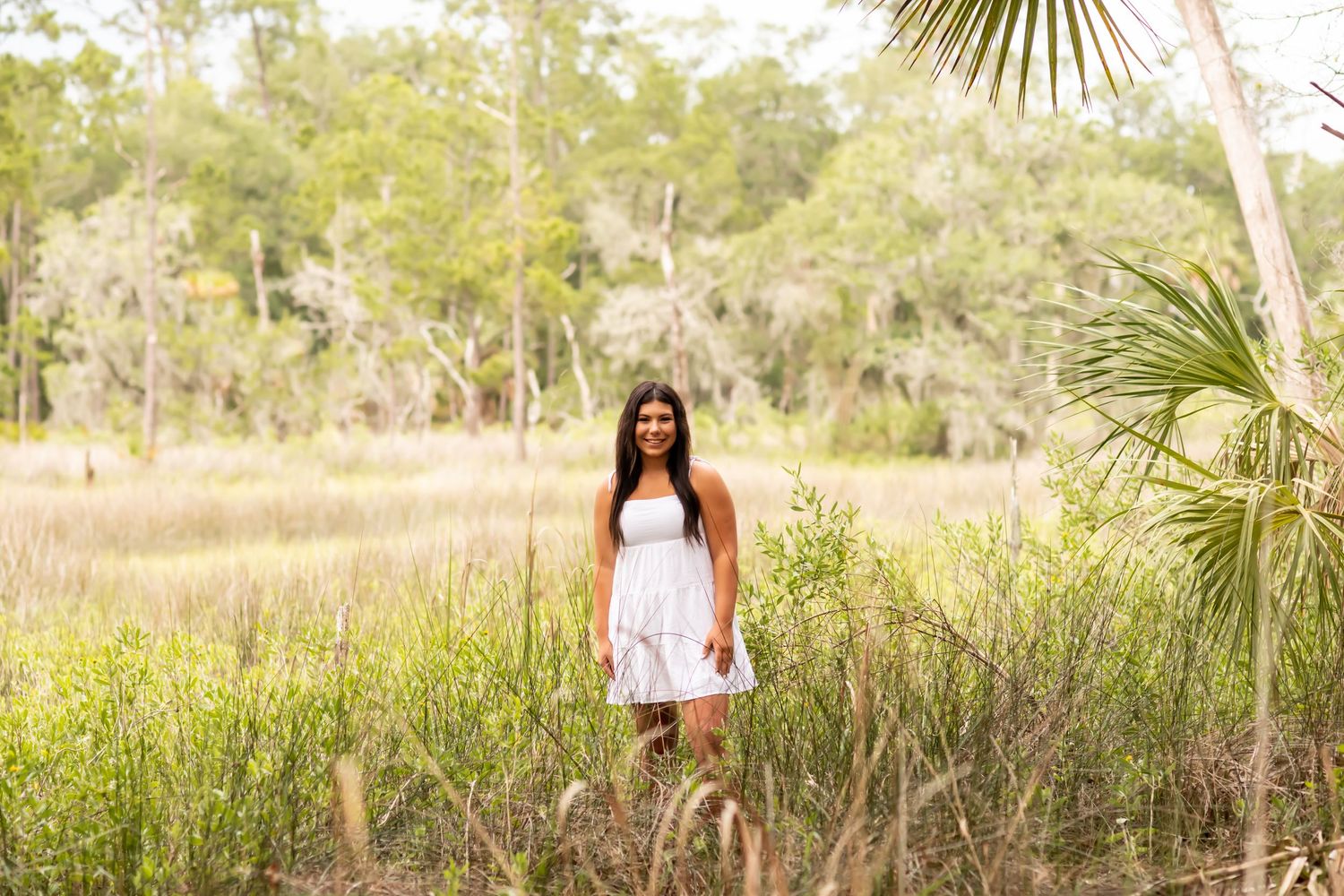 A person in a white dress stands in a field of tall grass with palm trees and woodlands in the background.