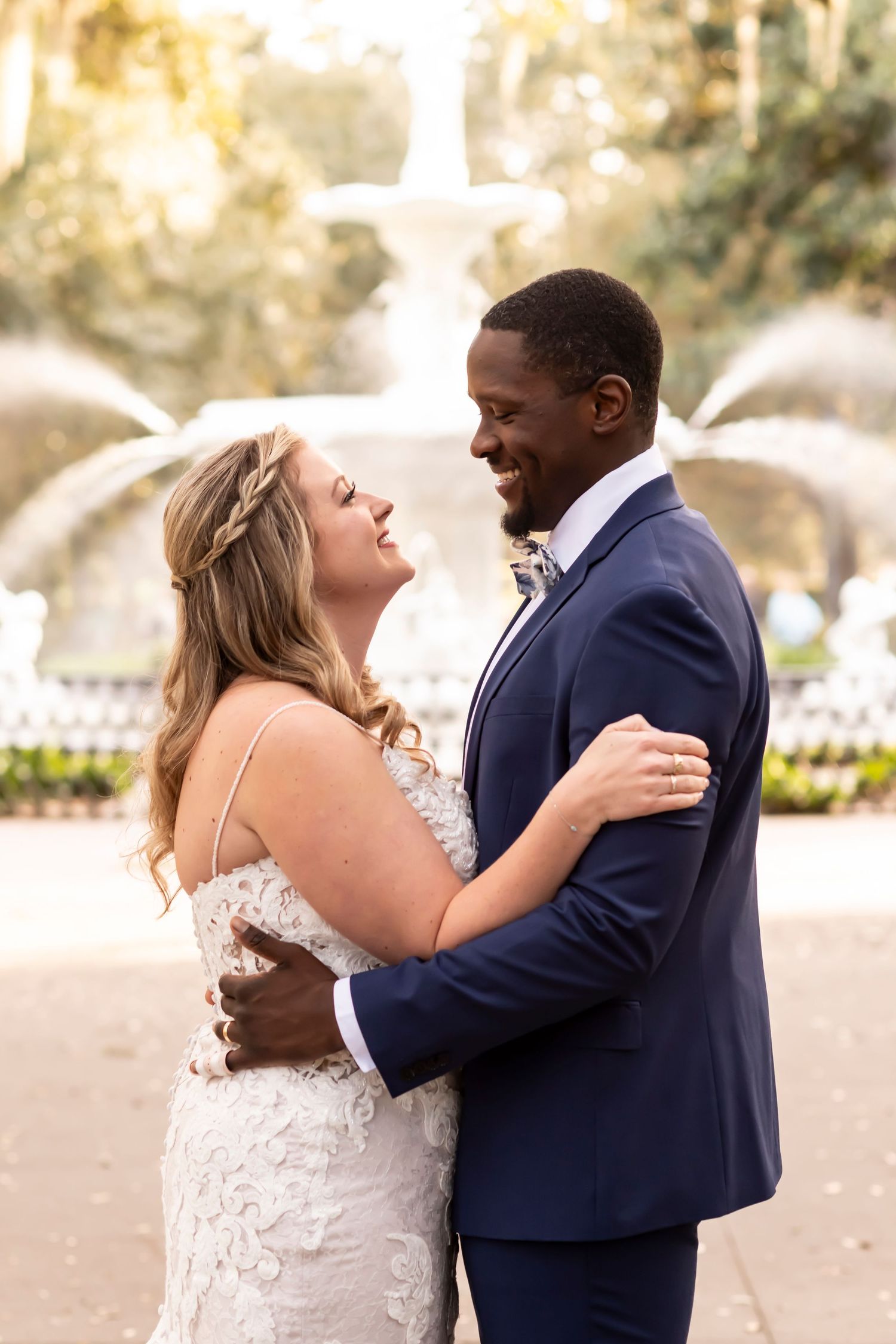 A couple embraces in front of an elegant fountain on their wedding day, wearing a lace gown and navy suit.