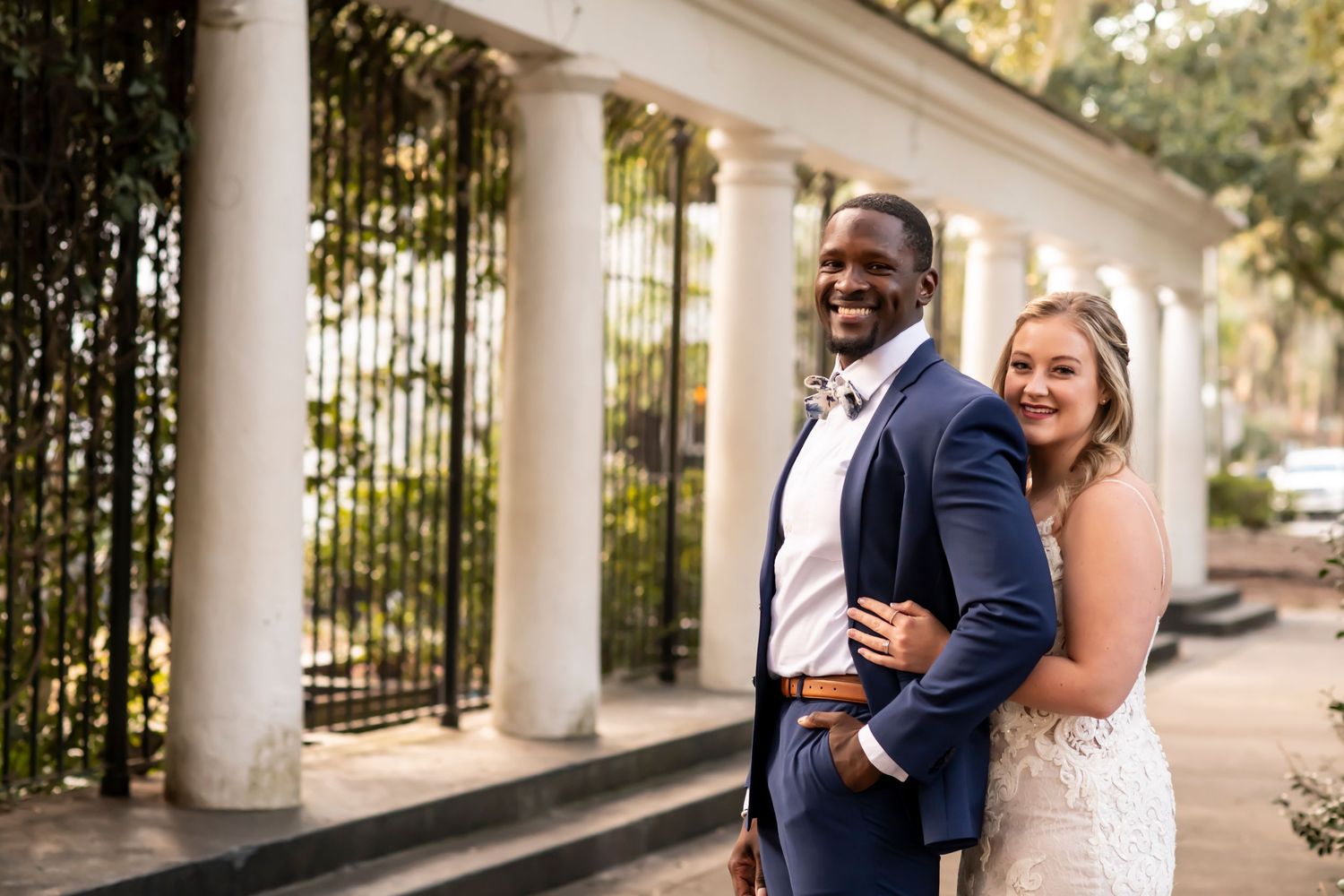 A cheerful couple laughing and embracing during their outdoor wedding photoshoot at a pillared walkway.