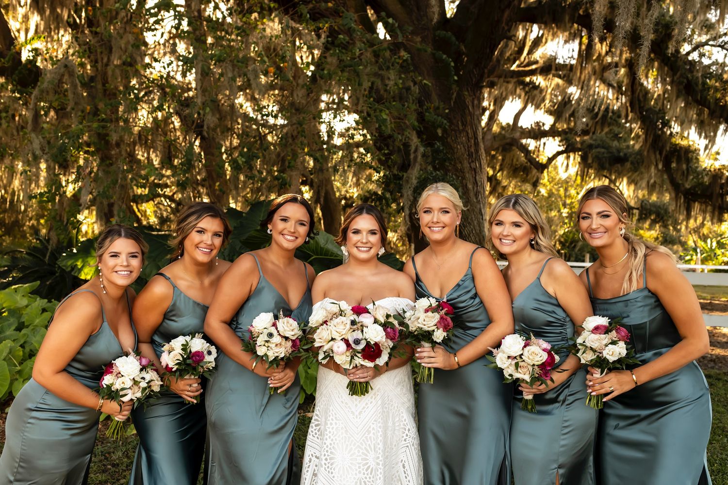 Bridesmaids in matching slate blue dresses holding colorful fall bouquets pose together outdoors under Spanish moss trees.