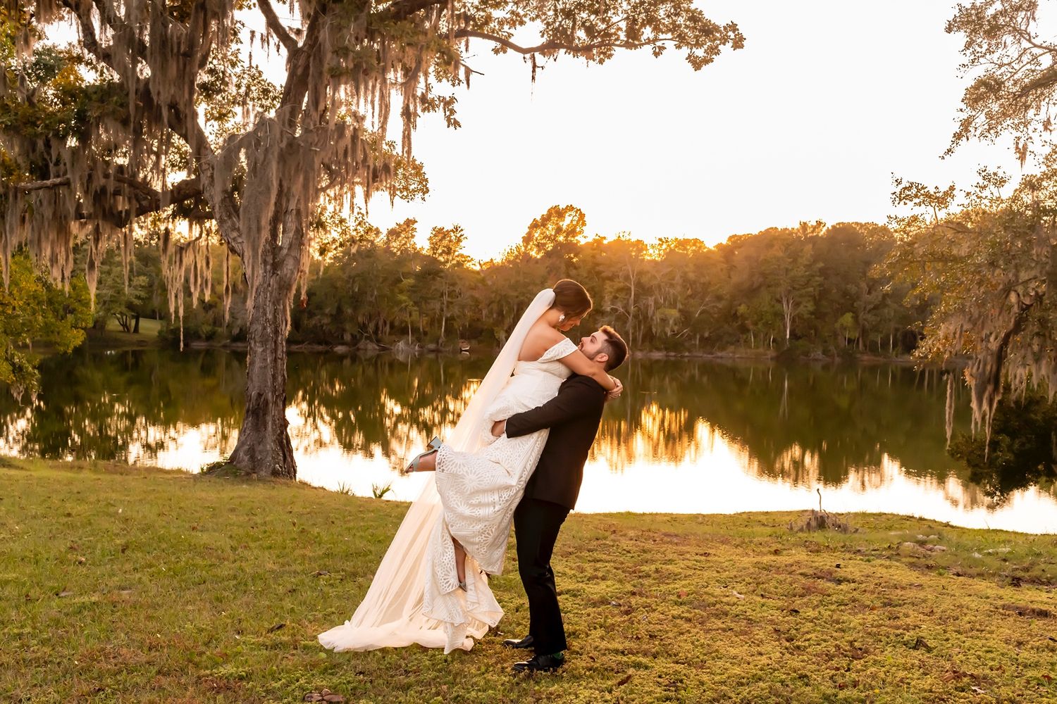 Couple embraces during golden hour sunset by a scenic lake with Spanish moss draped trees in the background.
