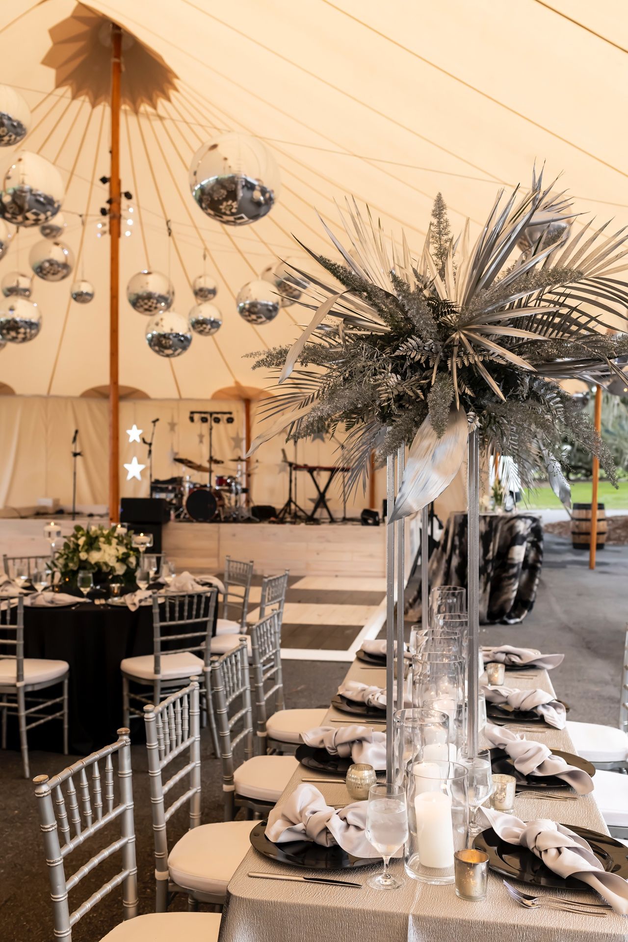 Elegant wedding tent interior with silver chiavari chairs, disco balls, and tall palm centerpieces in clear vases.