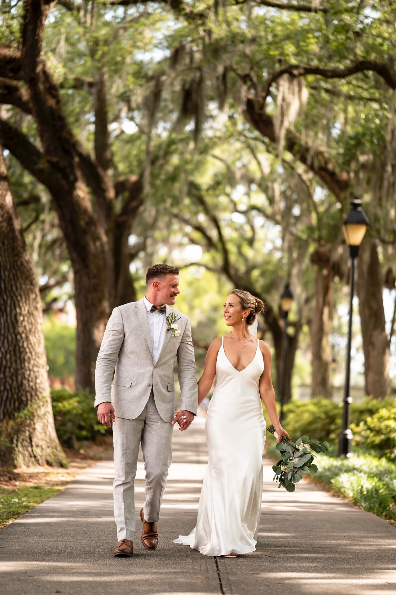 A couple walks hand in hand down a tree-lined path in Savannah Georgia, with Spanish moss draping the oak trees overhead.