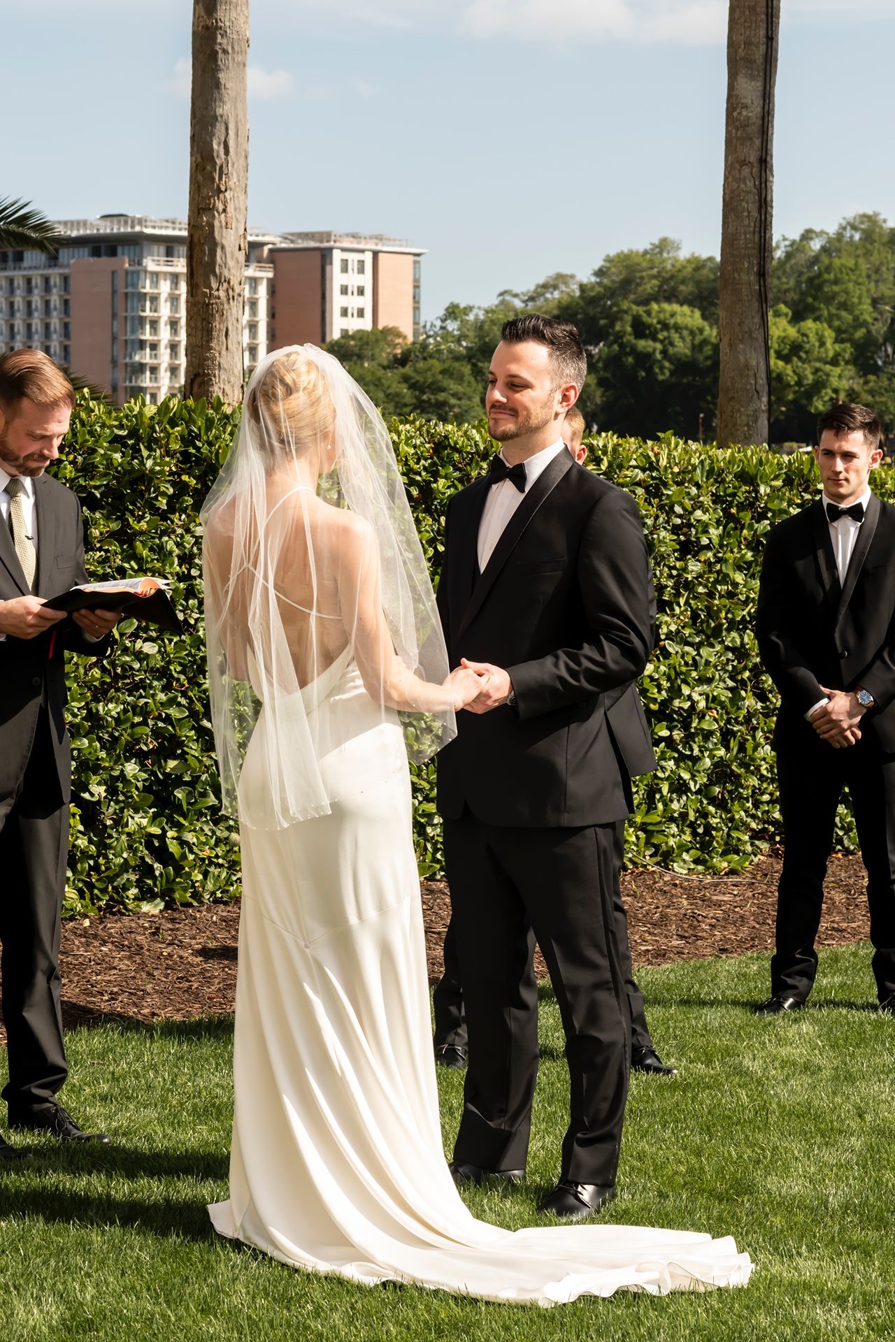 A bride and groom hold hands during an outdoor wedding ceremony with guests and a building in the background.