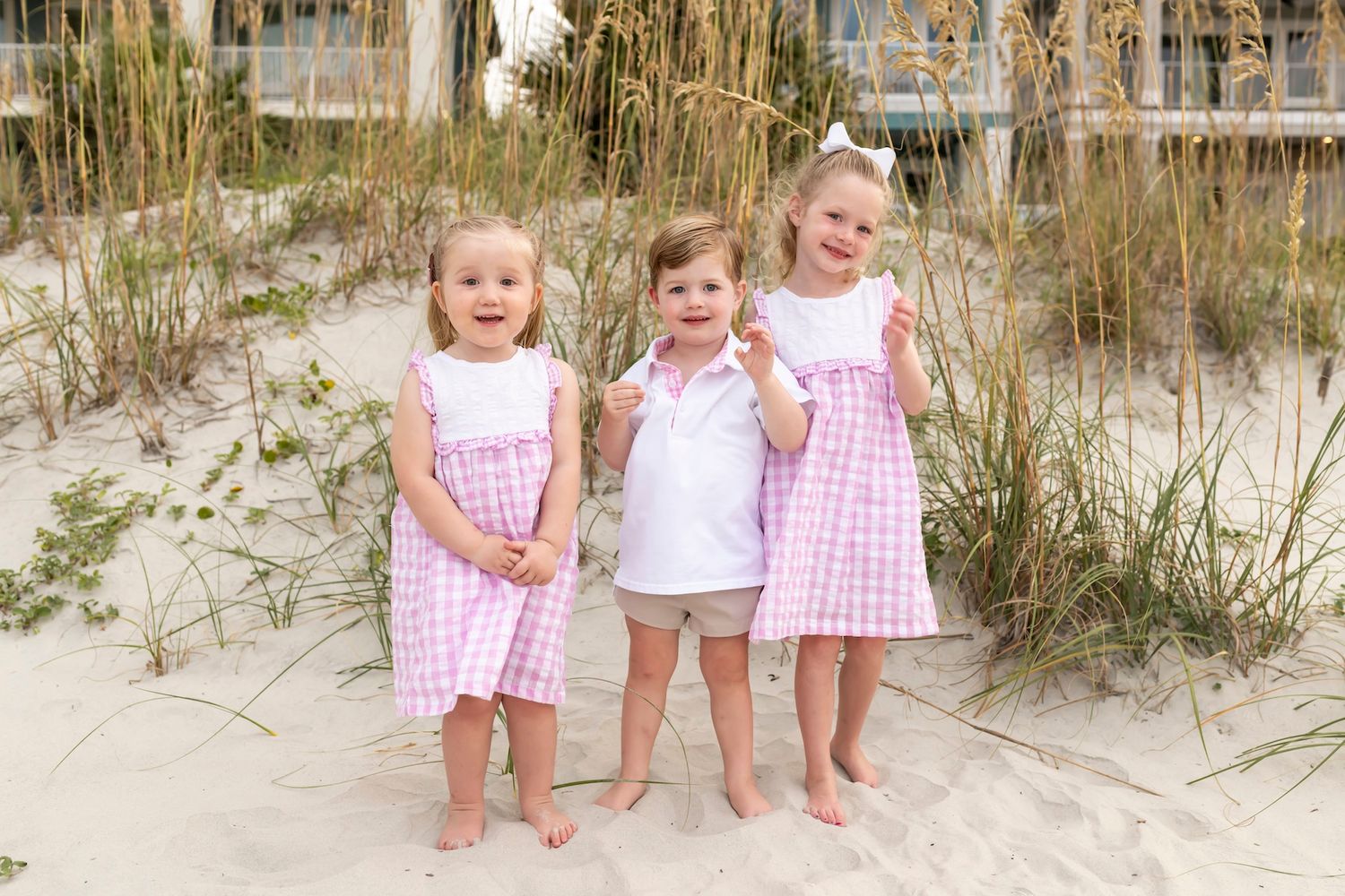 Four small children sitting on a sandy beach dune wearing white and pink outfits.