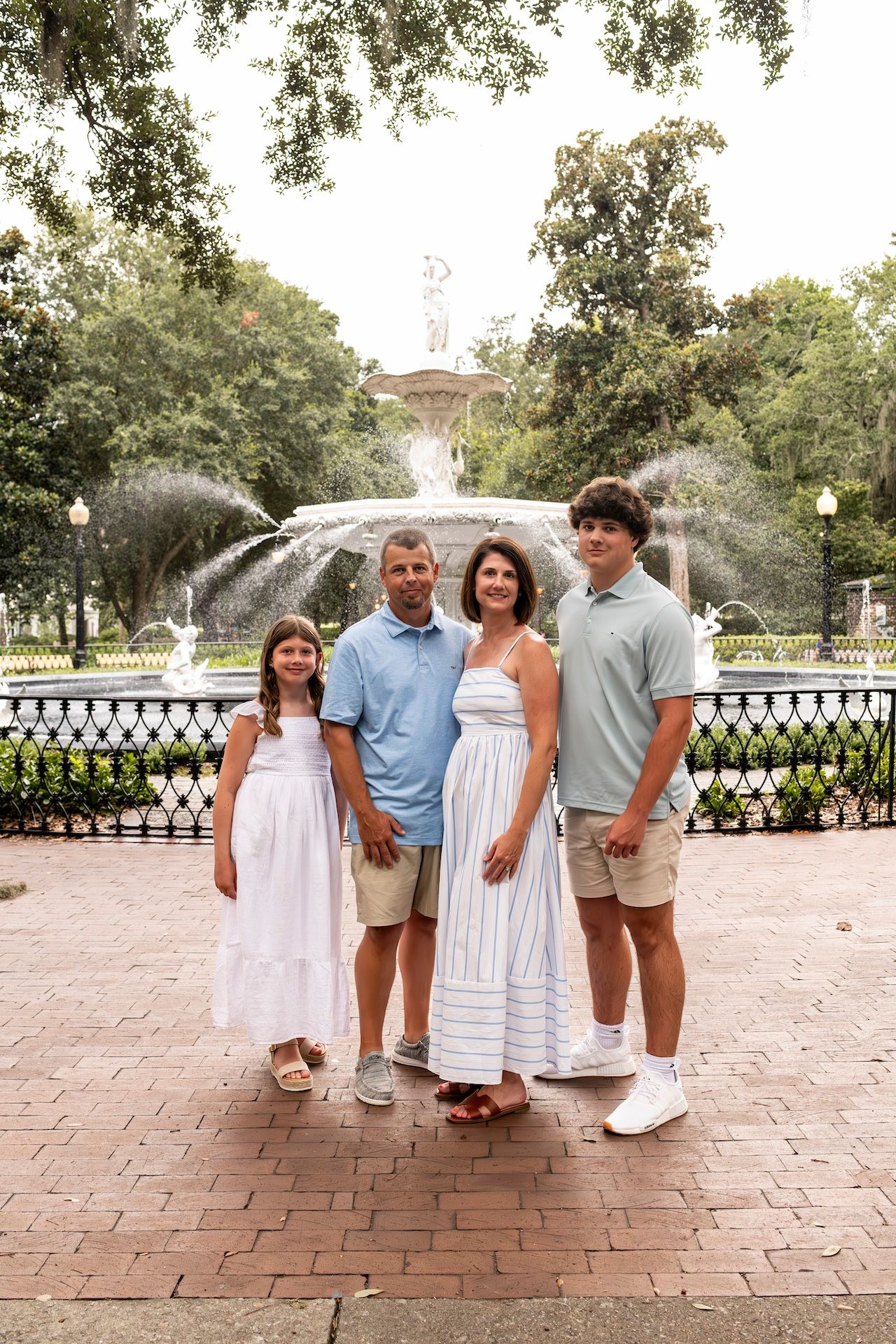 A family stands together in front of a fountain in a park with brick pavers and wrought iron fencing on a summer day.