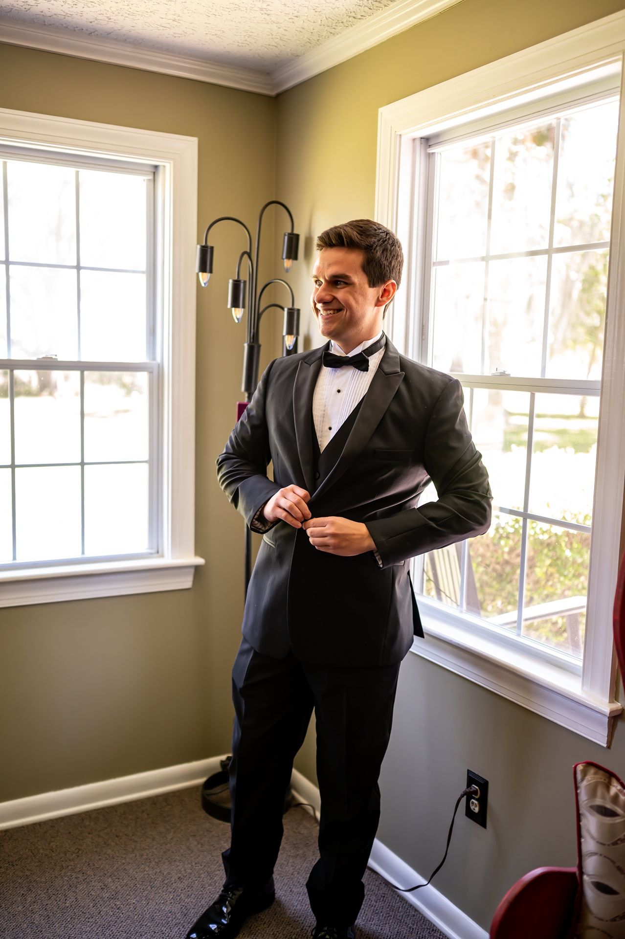 A groom in a classic black tuxedo and bow tie stands in a sunny room between two large windows.