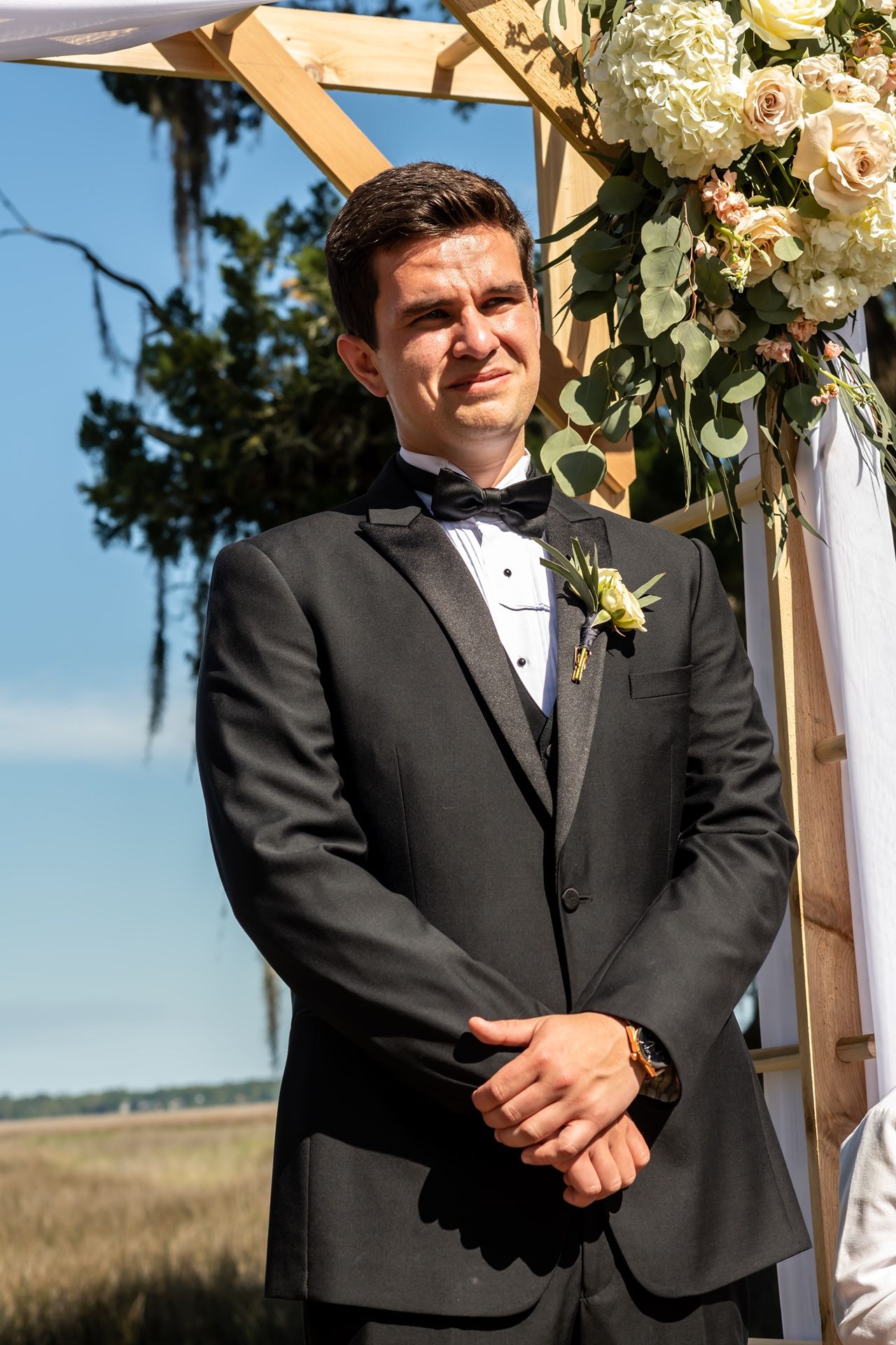 A groom in a black tuxedo and bowtie stands at an outdoor wedding ceremony arch decorated with white flowers.