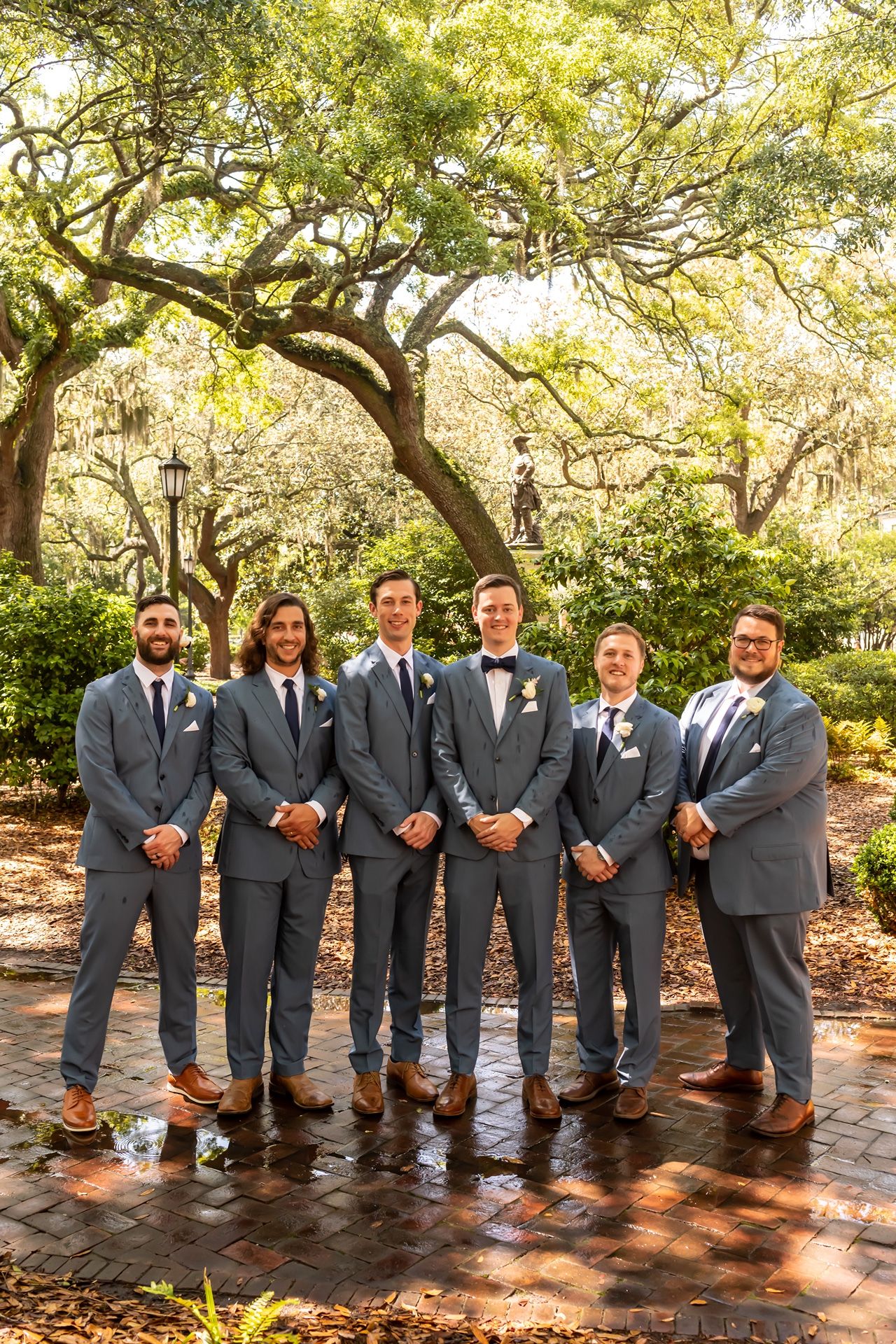 Wedding party in grey suits poses in sequence along brick pathway under sunlit trees in garden setting.