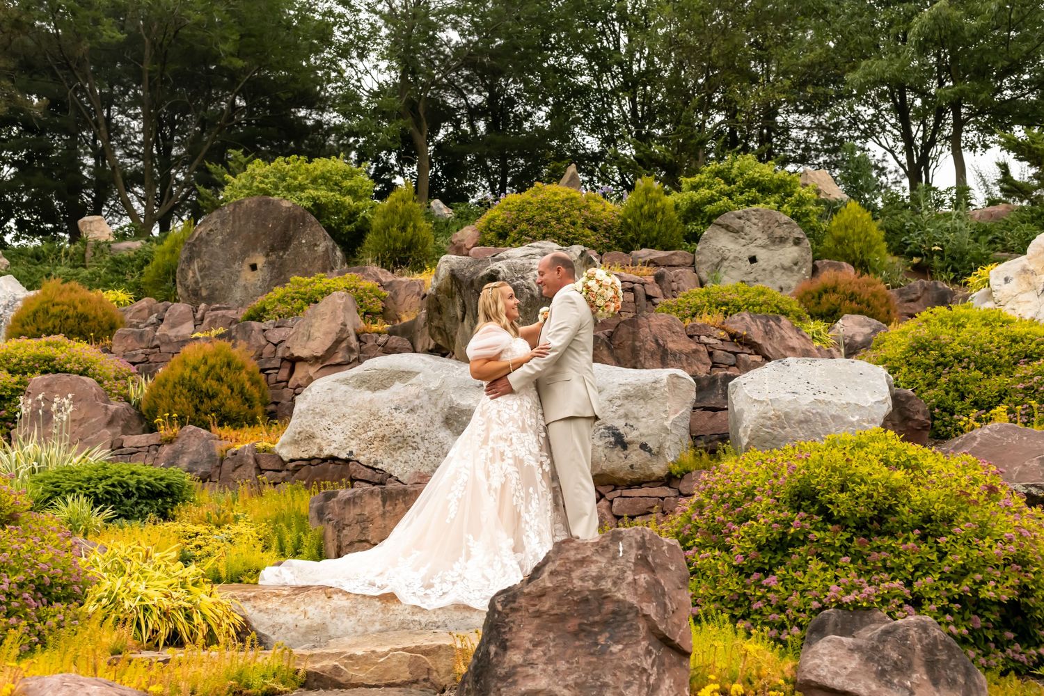 .Two people in wedding attire embrace on a landscaped hillside with large rocks.