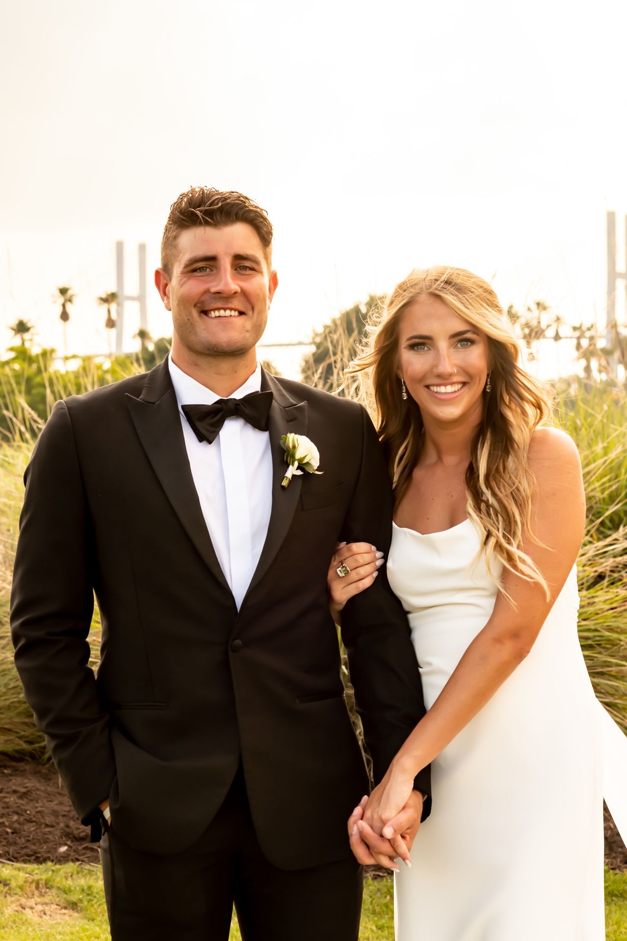 Wedding couple poses together outdoors at sunset in formal attire amidst palm trees and golden light.