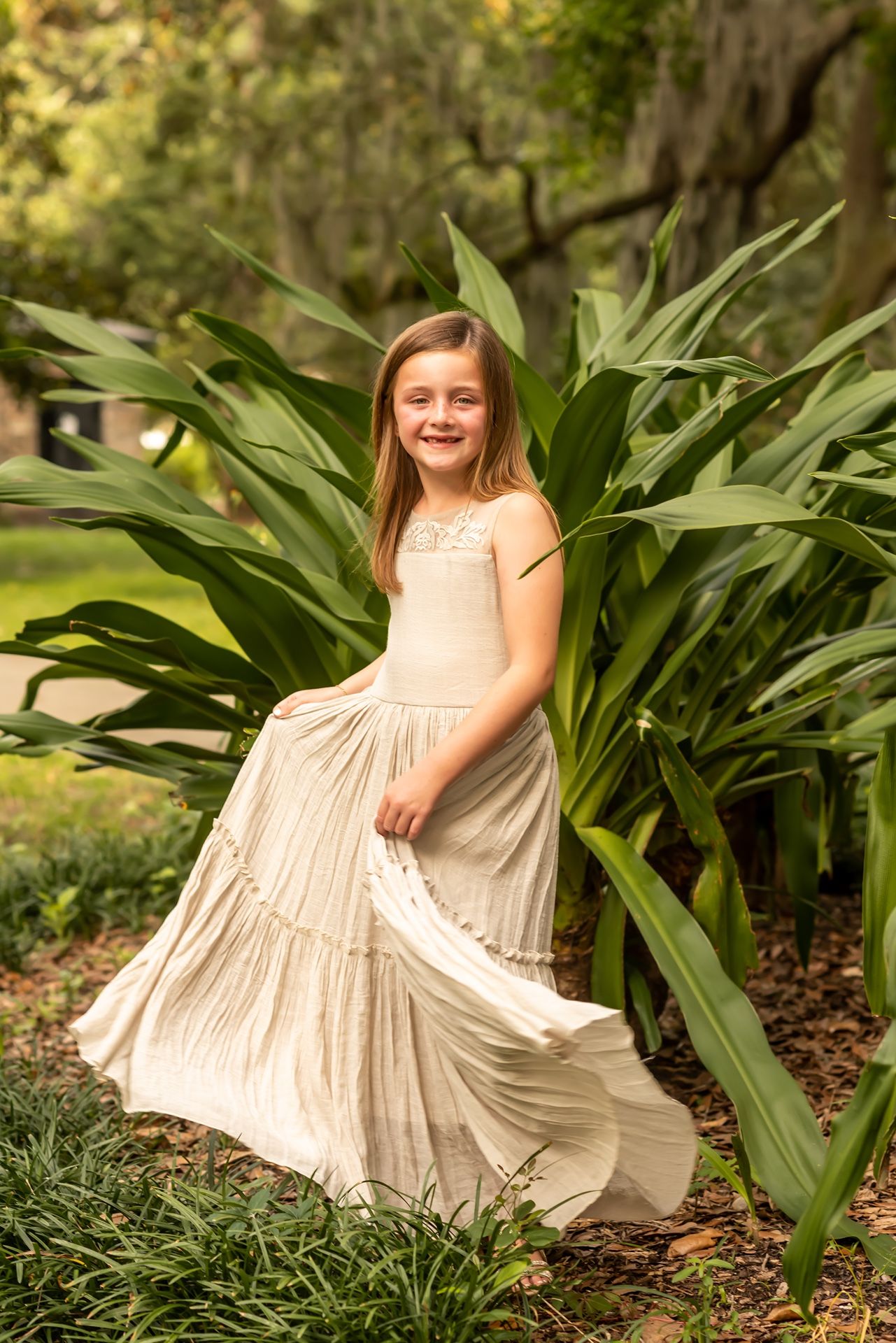 A cream colored tiered maxi dress flows elegantly against lush green foliage in a garden setting.
