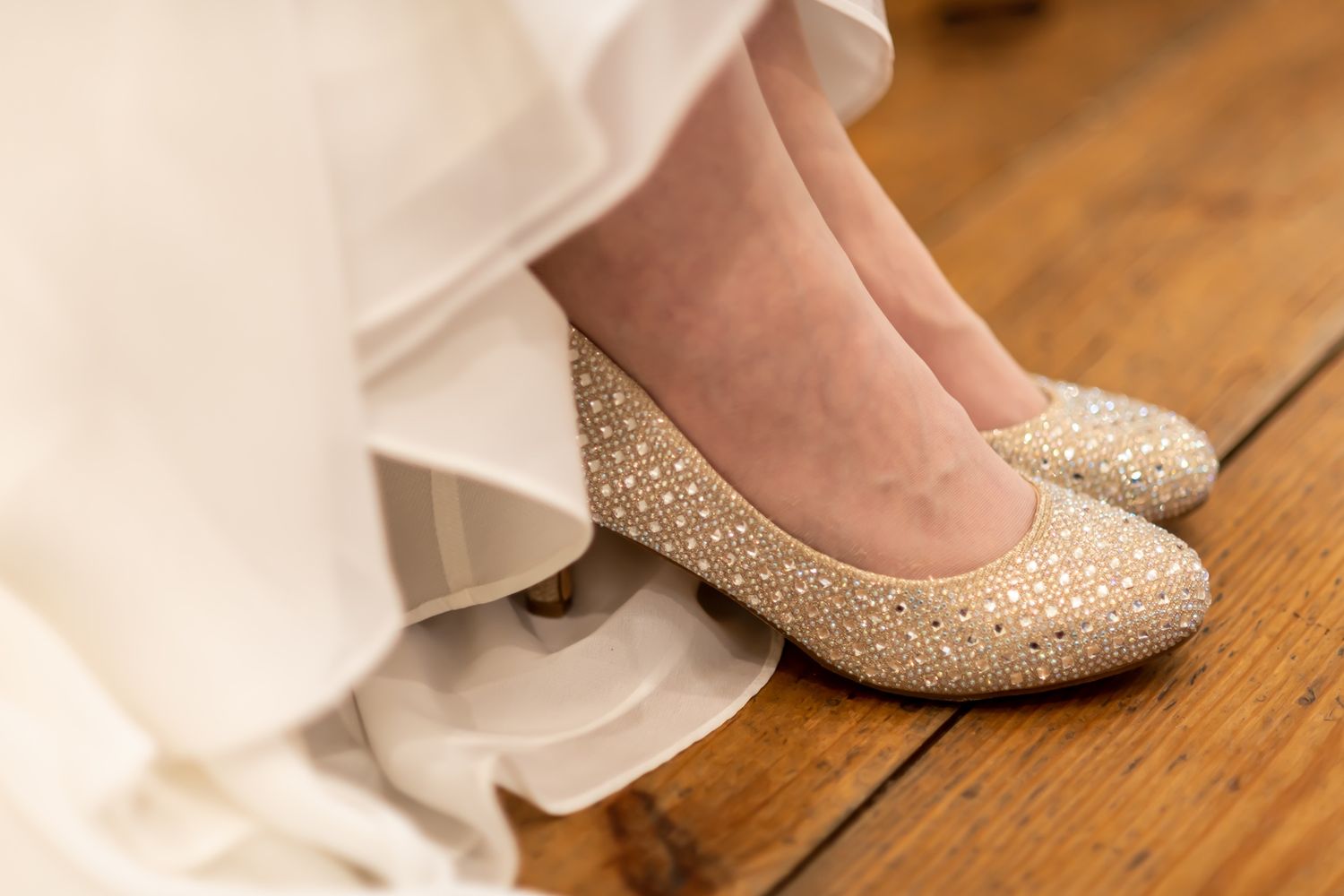 Glittery gold bridal shoes on a wooden floor with flowing white dress fabric.