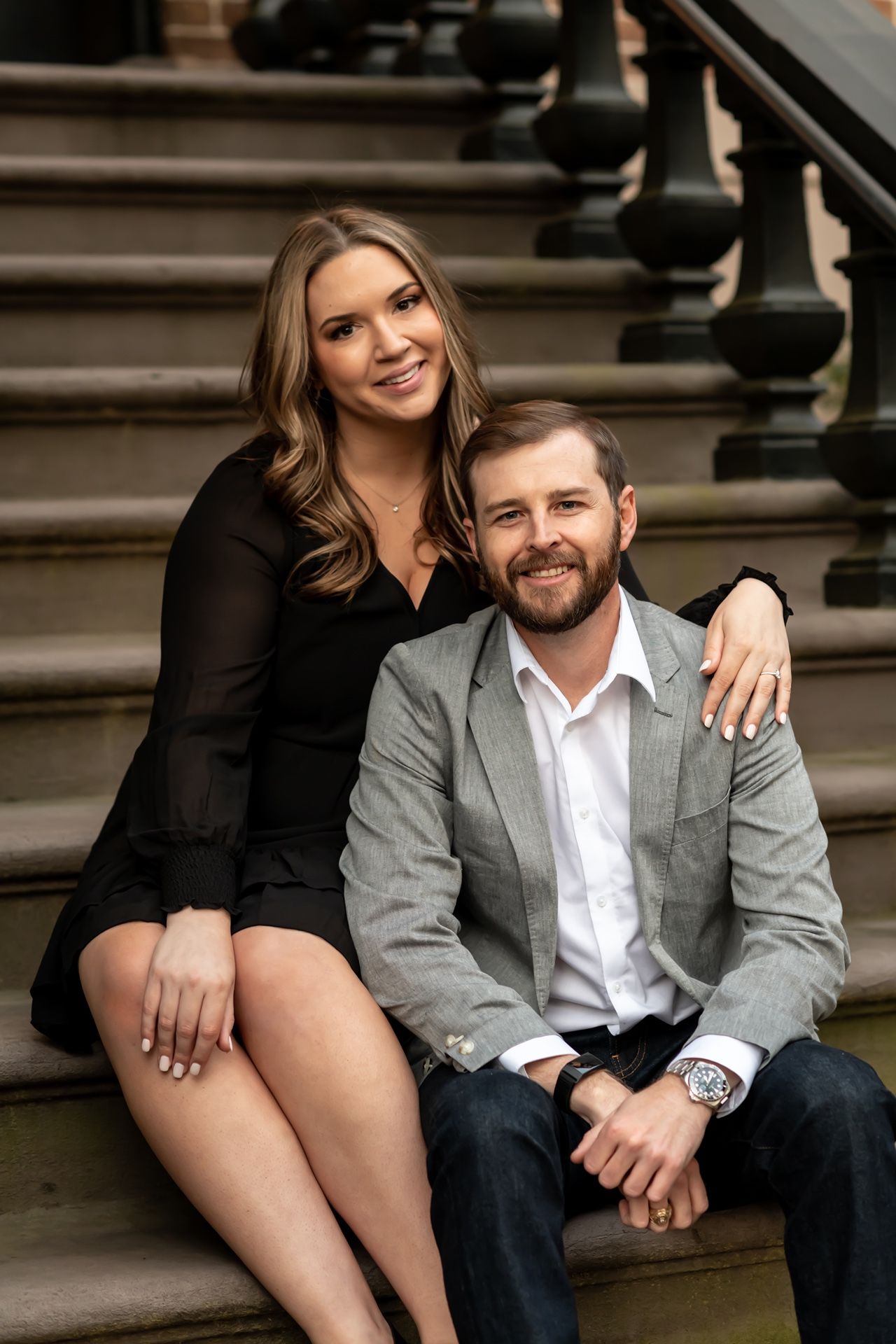 Couple sitting together on outdoor stone steps, dressed in black dress and gray blazer for engagement photos.