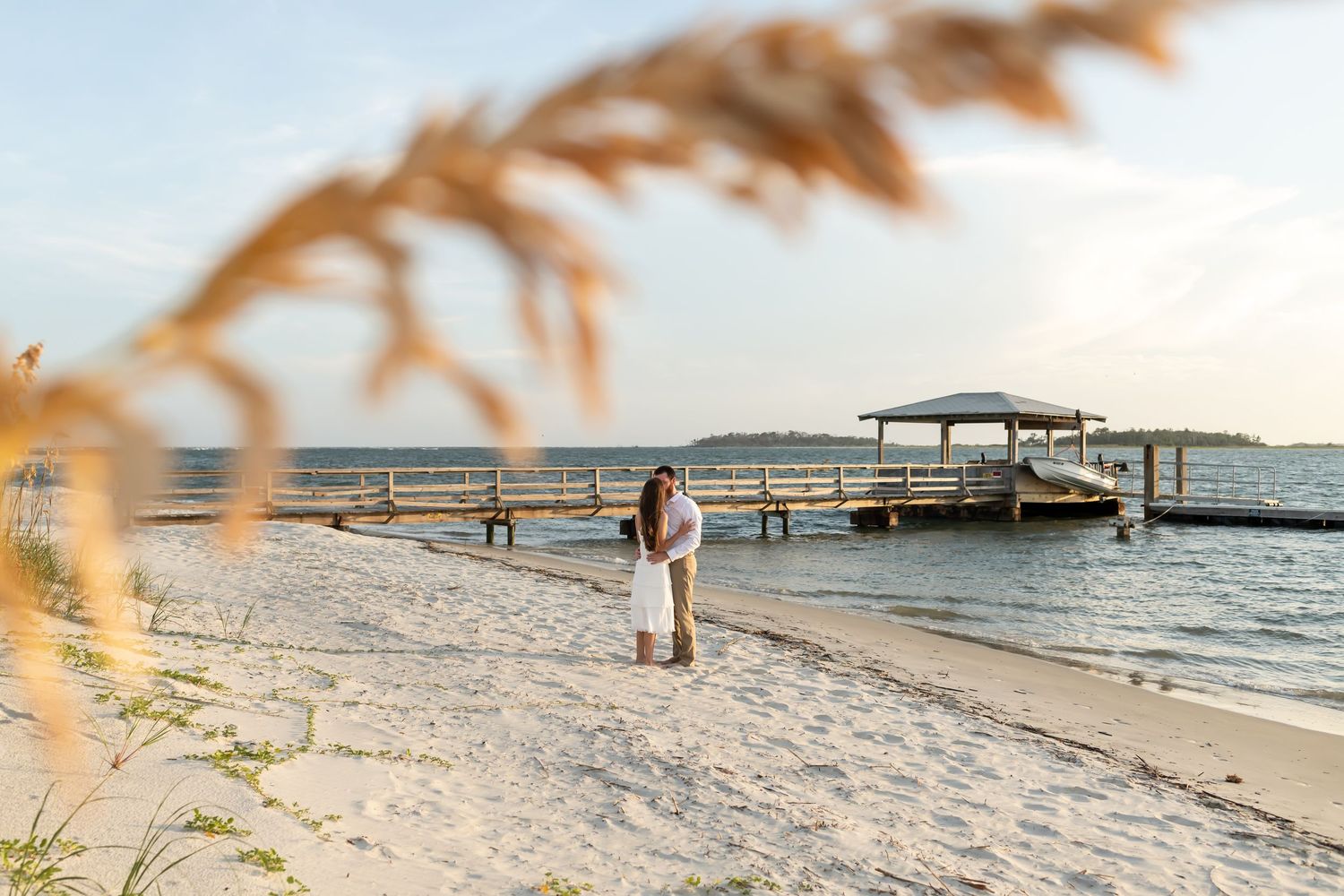 A couple stands on a sandy beach near a wooden pier during golden hour, with sea grass framing the scene.