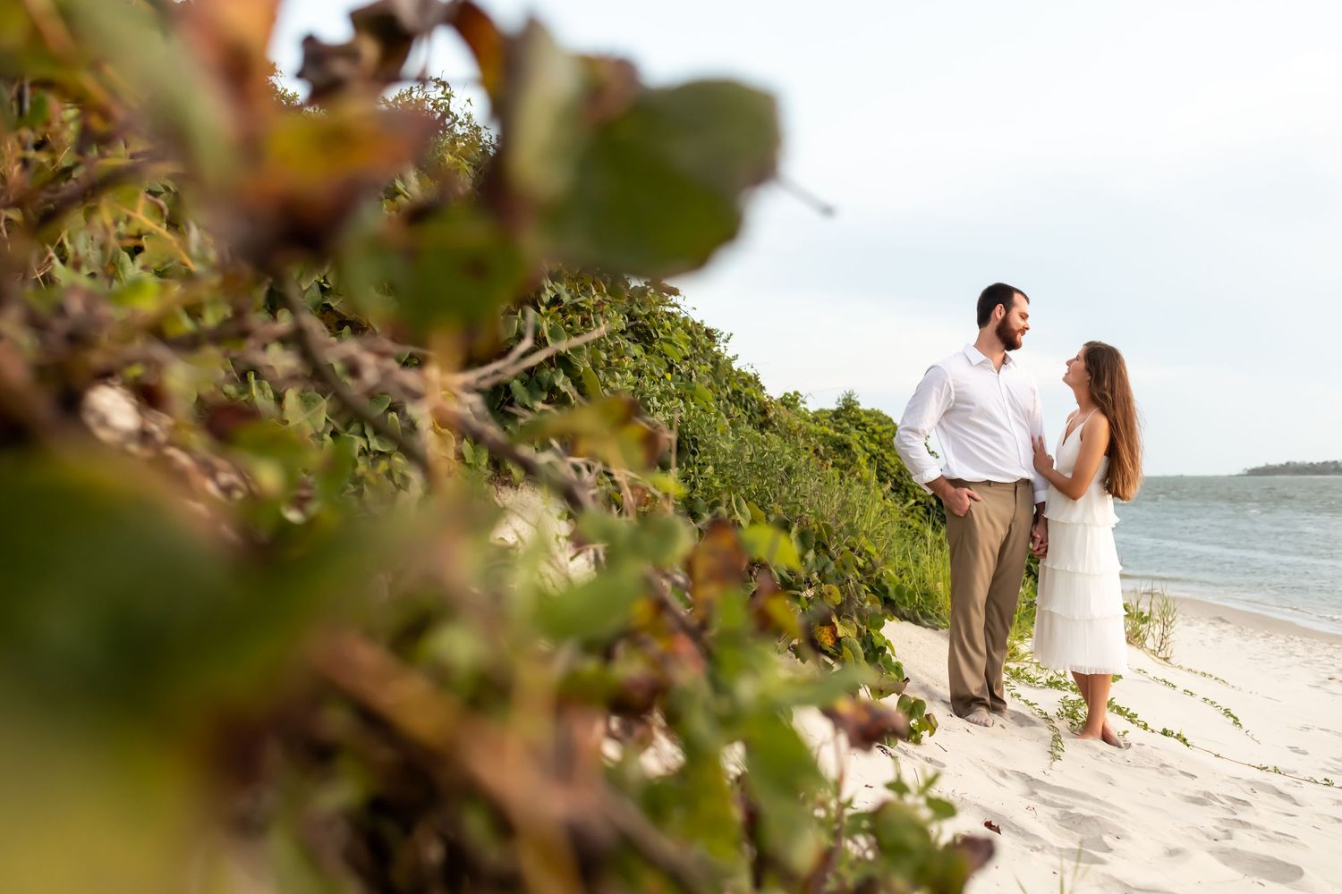 A couple stands together on a tropical beach with lush green foliage in the foreground and ocean in the background.